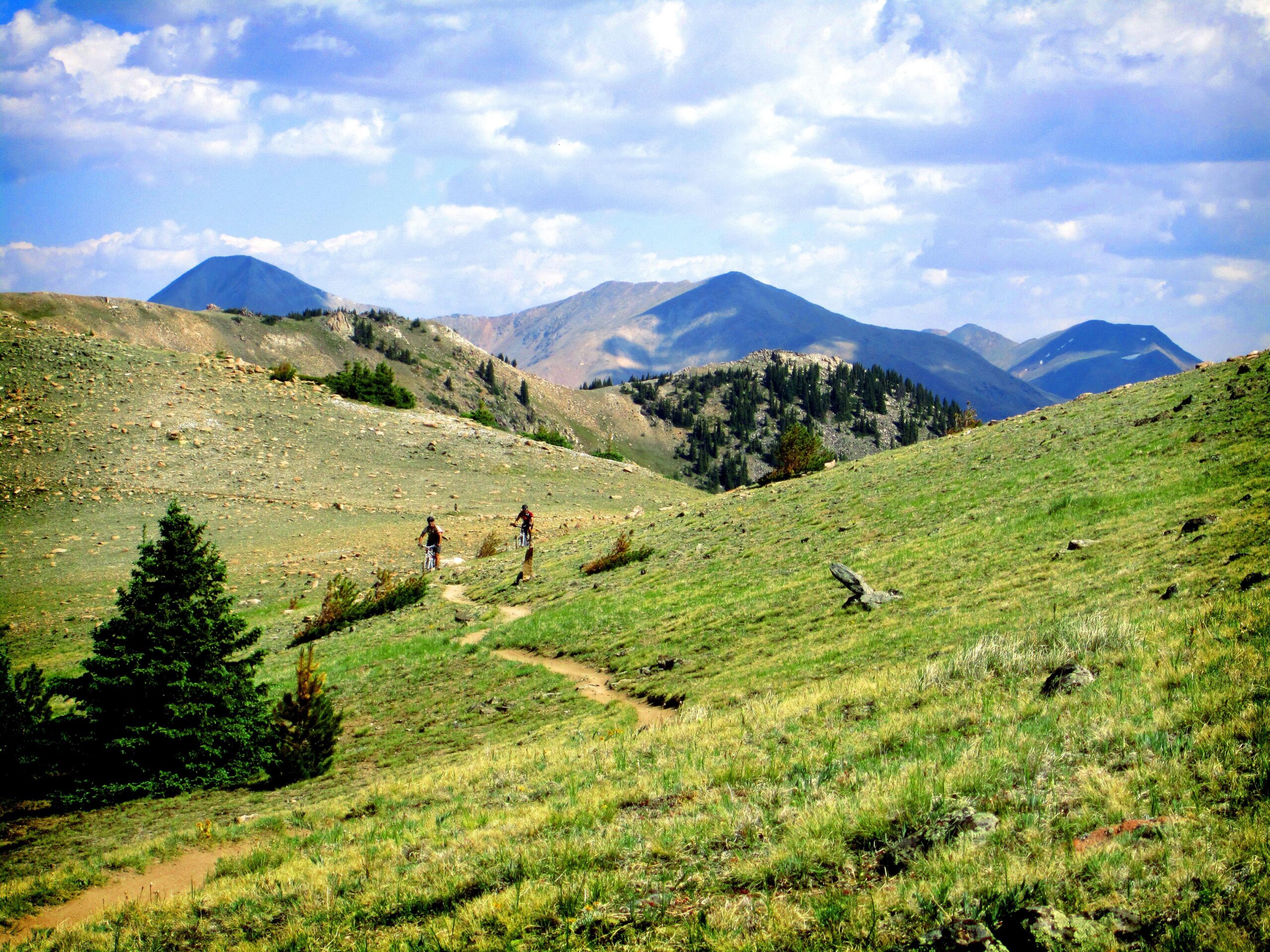 Two cyclists riding along a winding trail through a green, hilly landscape with rocky outcrops and trees, set against a backdrop of distant mountains under a partly cloudy sky. Monarch Crest Trail mountain bike trail.