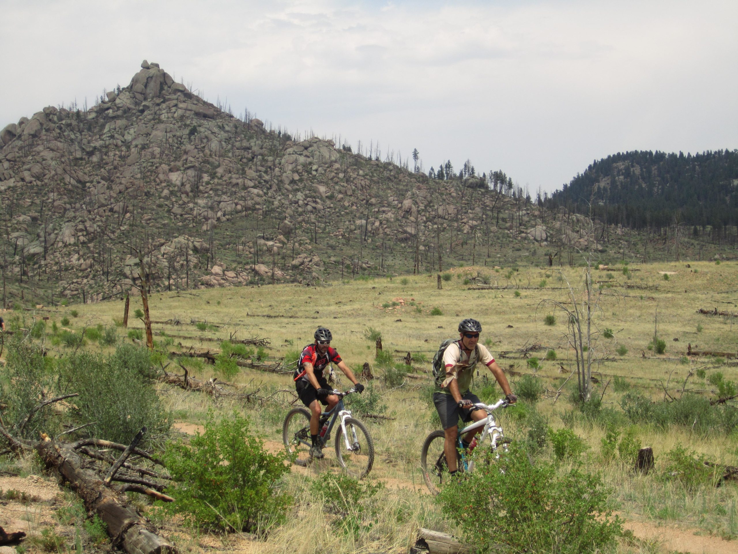 Two mountain bikers ride through a rocky, natural landscape with sparse vegetation and patches of green. In the background, a prominent rocky hill rises against a cloudy sky, while charred tree stumps and fallen logs are scattered throughout the scene. Buffalo Creek mountain bike trail.