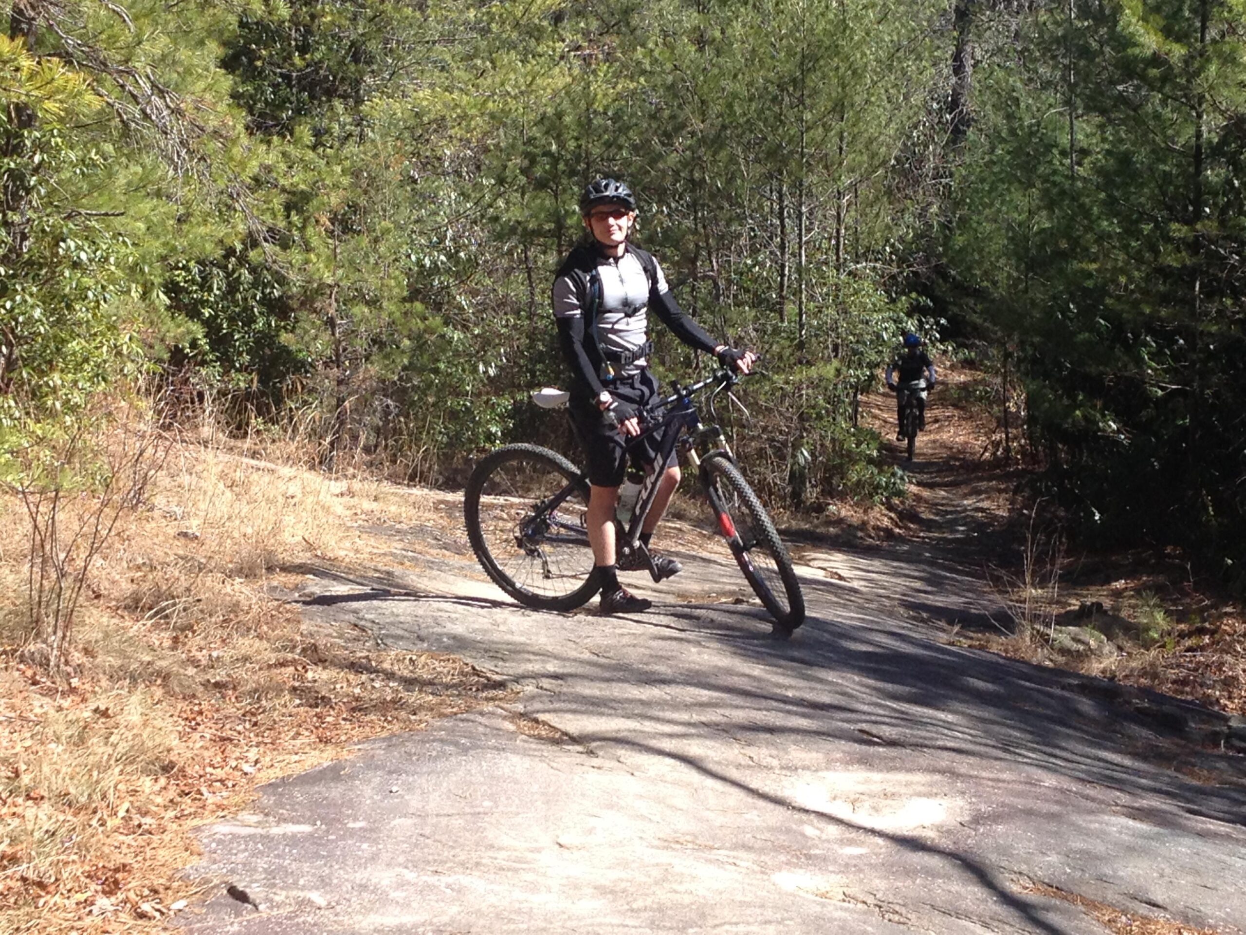 A mountain biker in cycling attire stands beside his bike on a rocky trail surrounded by trees. In the background, another cyclist is riding along the path. The scene captures a sunny day in a natural setting, emphasizing outdoor activities and mountain biking. Cedar Rock Trail #16 mountain bike trail.