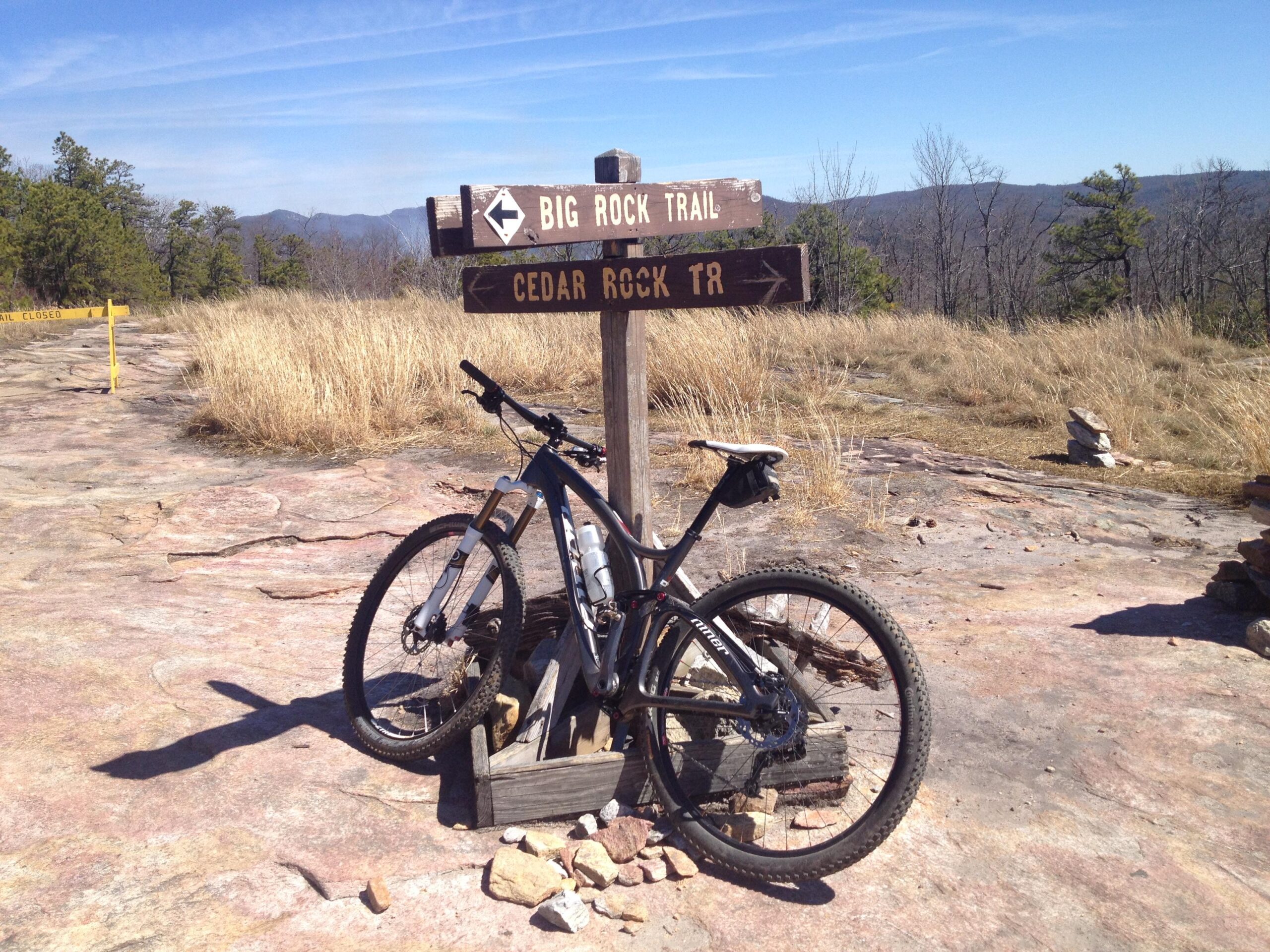 A mountain bike leaning against a wooden trail signpost at a rocky trail intersection. The sign indicates directions for "Big Rock Trail" and "Cedar Rock TR." The background features a landscape of grassy areas and distant mountains under a clear blue sky. A "Trail Closed" warning sign is visible in the distance. Cedar Rock Trail #16 mountain bike trail.