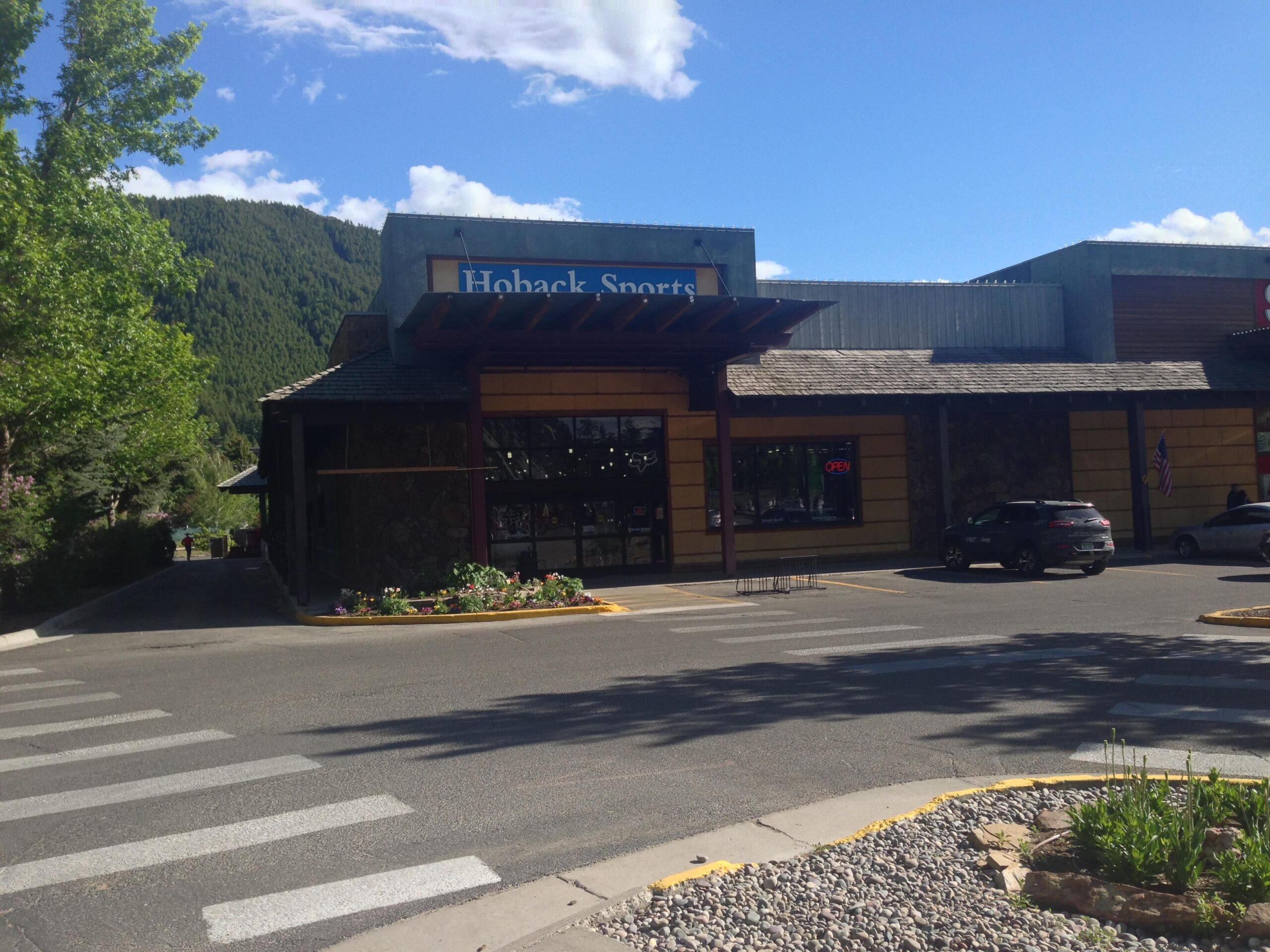 A storefront of Hoback Sports, featuring a large glass window displaying the "OPEN" sign. A paved parking lot in front includes a parked vehicle and a crosswalk. Surrounding the store are trees and landscaped flowerbeds, with a backdrop of green mountains under a blue sky with some clouds.