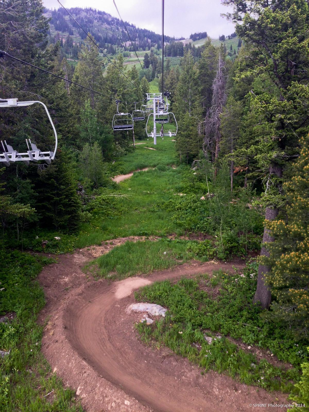 A view of a ski lift moving through a forested area, with several empty chairs hanging from cables above. Below, a dirt path winds through lush green grass and trees, leading into a natural landscape of rolling hills and mountain terrain in the background. Jackson Hole Bike Park mountain bike trail.