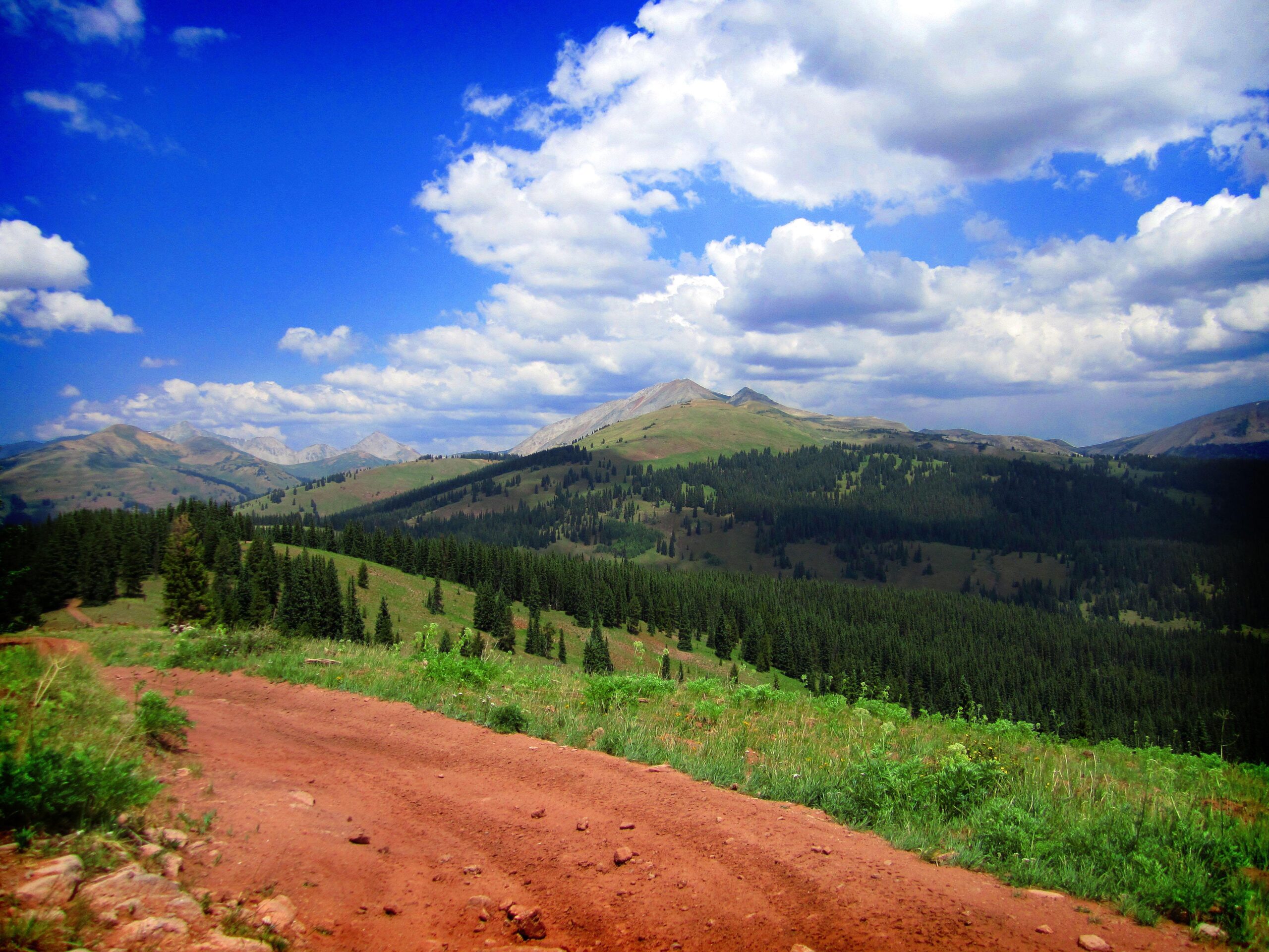 A scenic view of a mountainous landscape featuring rolling green hills, dense forests of evergreen trees, and distant peaks under a bright blue sky with fluffy white clouds. A dirt path winds through the foreground, inviting exploration of the natural surroundings. Reno / Flag / Bear / Deadman Loop mountain bike trail.