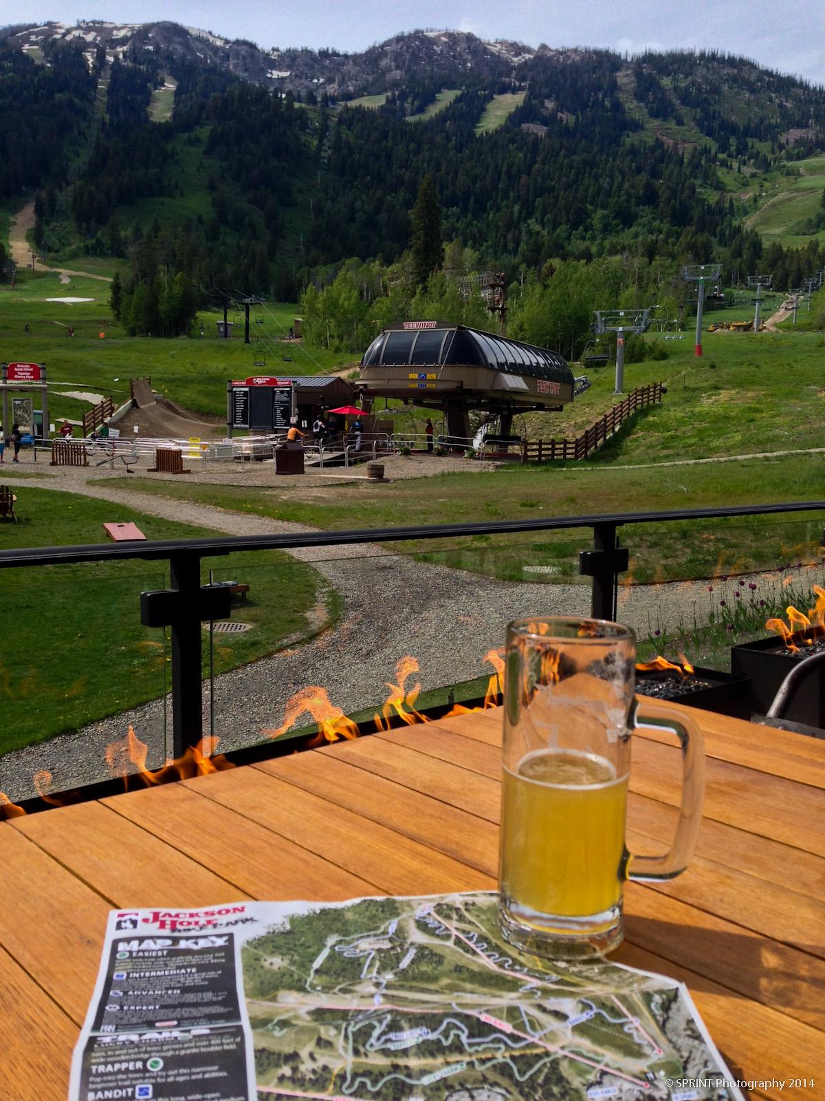 A scenic view from a balcony featuring a wooden table with a glass of beer and a trail map. In the background, ski lifts are visible against a lush green hillside, with distant mountains partially covered in snow. The setting suggests a relaxed outdoor atmosphere in a mountain resort. Jackson Hole Bike Park mountain bike trail.