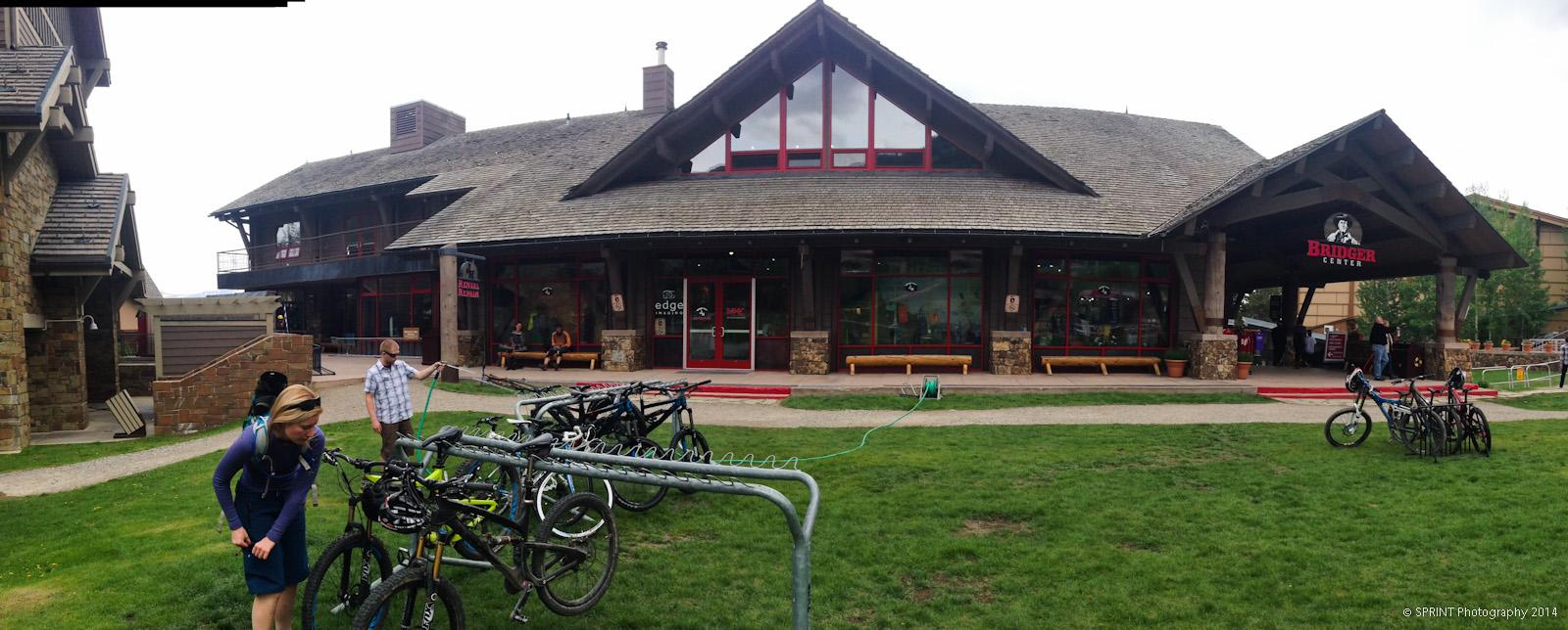 A modern building with a wooden exterior and large windows, labeled "Bridger Center." In the foreground, several bicycles are parked in a rack, and a woman is preparing her bike while a man stands nearby holding a hose. The lawn is green, and there are benches along the building