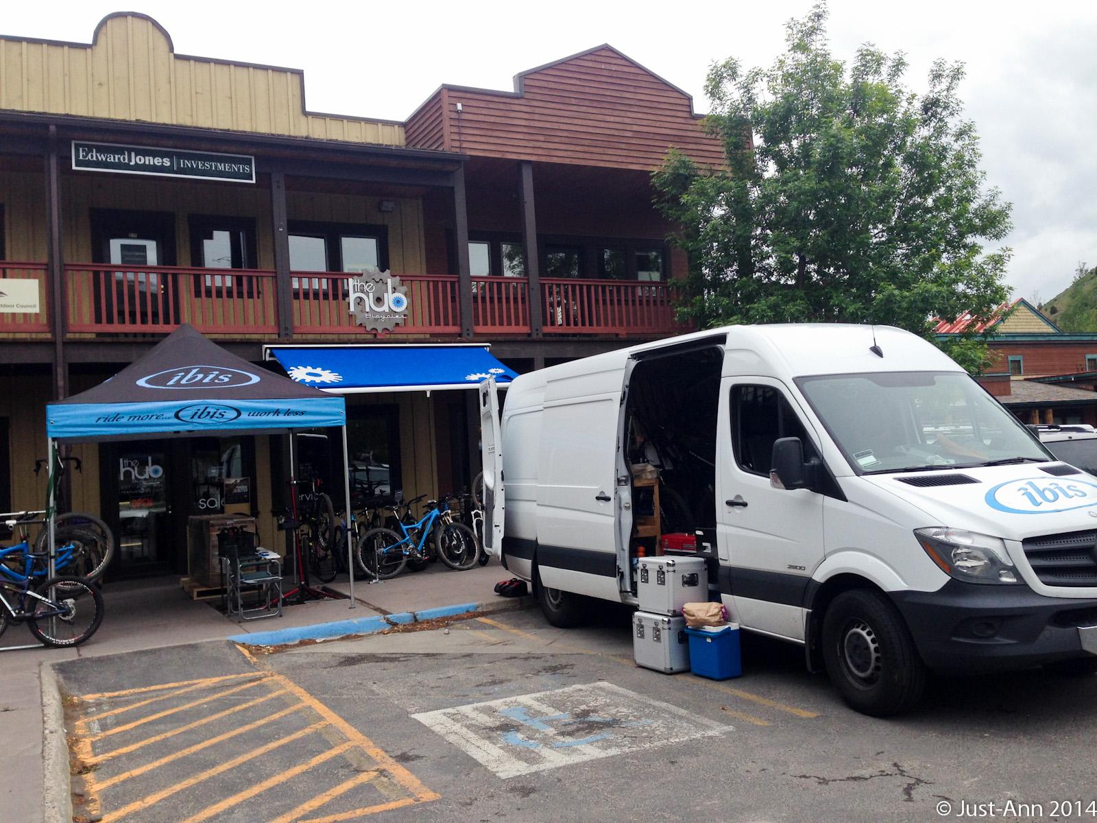 A white van parked outside a bike shop with an open side door revealing equipment. A blue canopy featuring the Ibis logo is set up in front of the shop, which has several bicycles displayed outside. The shop has a wooden fa&ccedil;ade and features a sign for Edward Jones Investments above. Trees and a cloudy sky can be seen in the background.