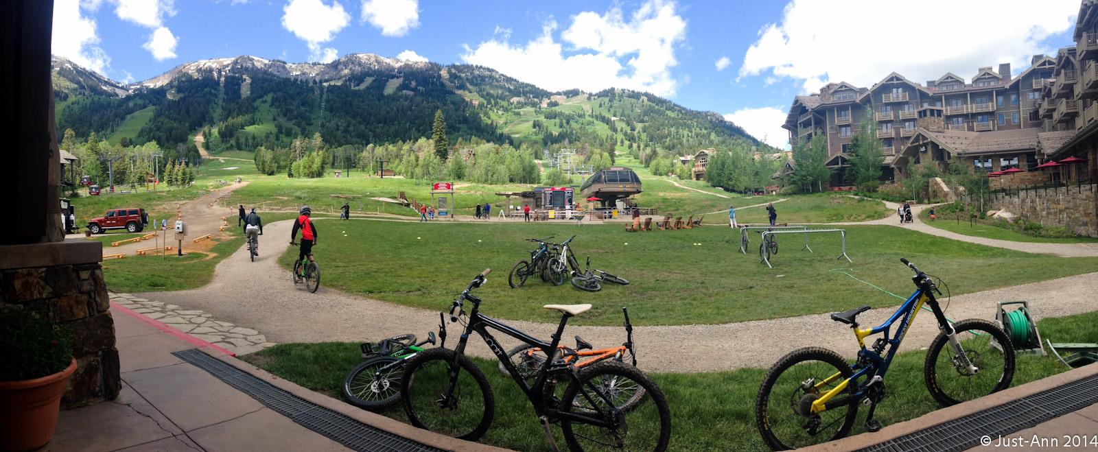 A panoramic view of a mountainous landscape featuring a green valley with a gravel path, where several cyclists are riding. In the foreground, a variety of mountain bikes are parked, while various people are enjoying the outdoor atmosphere. The backdrop showcases snow-capped mountains under a blue sky with scattered clouds. A lodge and seating area are visible on the right side of the image. Jackson Hole Bike Park mountain bike trail.