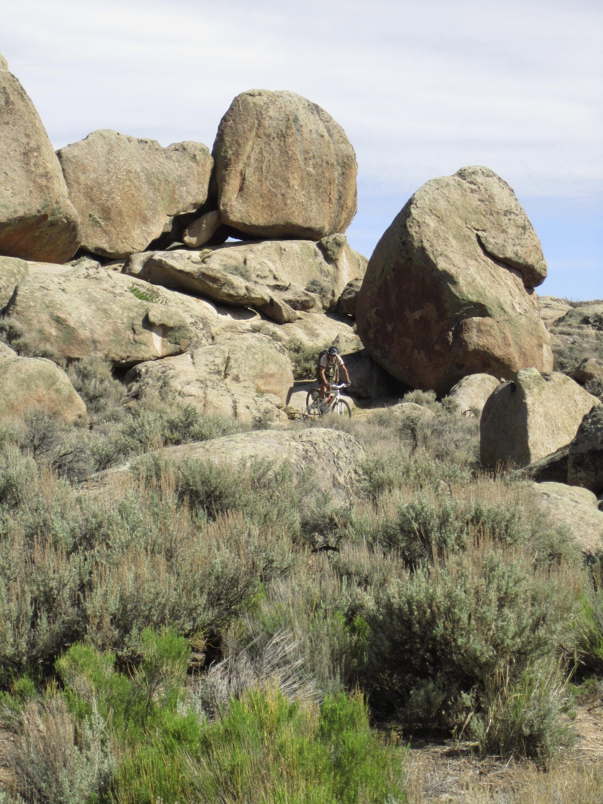A mountain biker navigating a rocky terrain surrounded by large boulders and sparse vegetation under a partly cloudy sky. Hartman Rocks mountain bike trail.