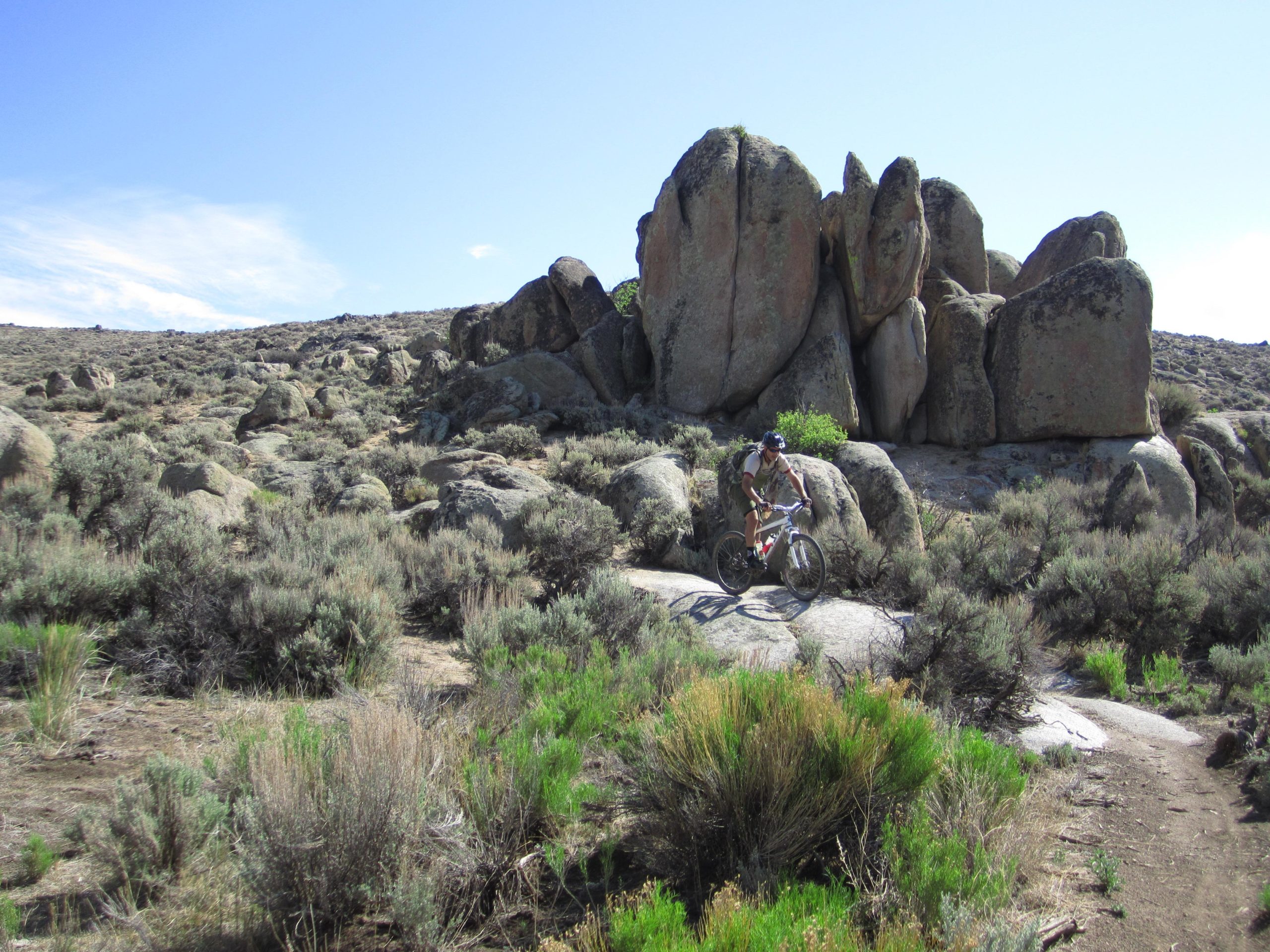 A mountain biker navigates rocky terrain surrounded by shrubs and boulders under a clear blue sky. Hartman Rocks mountain bike trail.