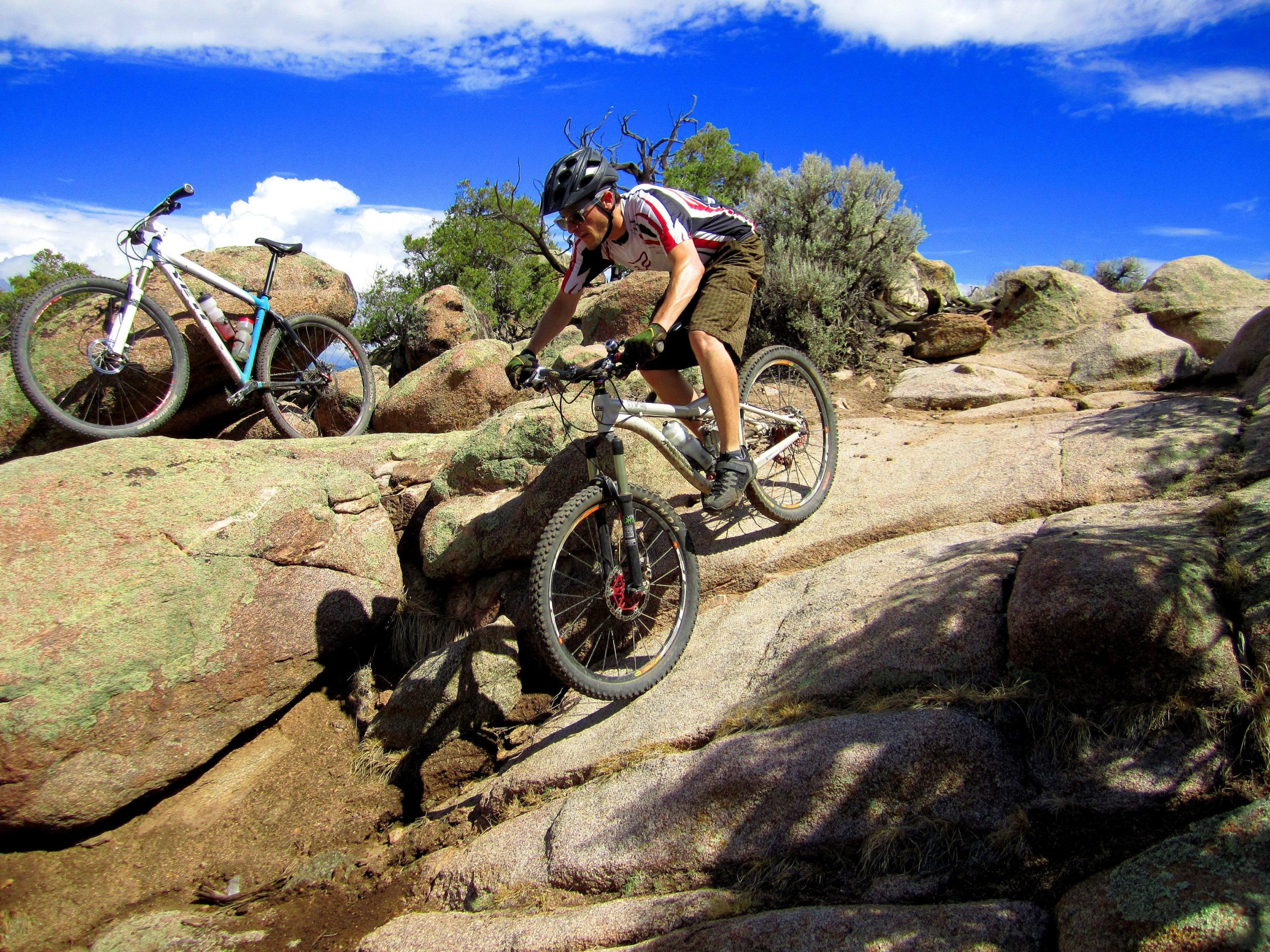 A person in a helmet riding a mountain bike over rocky terrain, with a second bike parked on the rocks nearby. The scene includes blue skies and scattered clouds, surrounded by vegetation and boulders. Hartman Rocks mountain bike trail.