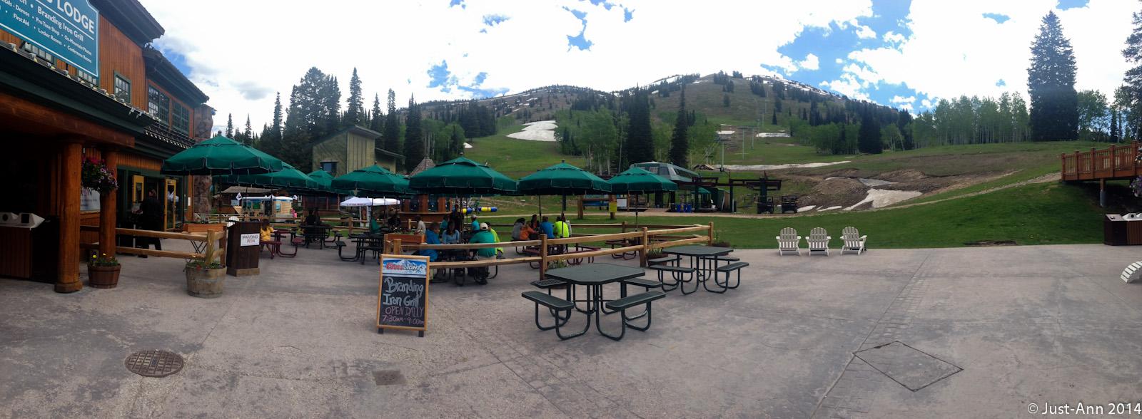 A scenic view of a mountain lodge area with outdoor seating beneath green umbrellas. Several picnic tables are arranged on a paved surface, and a sign advertising "Branding Iron Grill" can be seen. In the background, a hillside is visible with some remnants of snow and tall trees, under a partly cloudy sky. The atmosphere appears inviting and perfect for outdoor dining or relaxation. Grand Targhee Bike Park mountain bike trail.