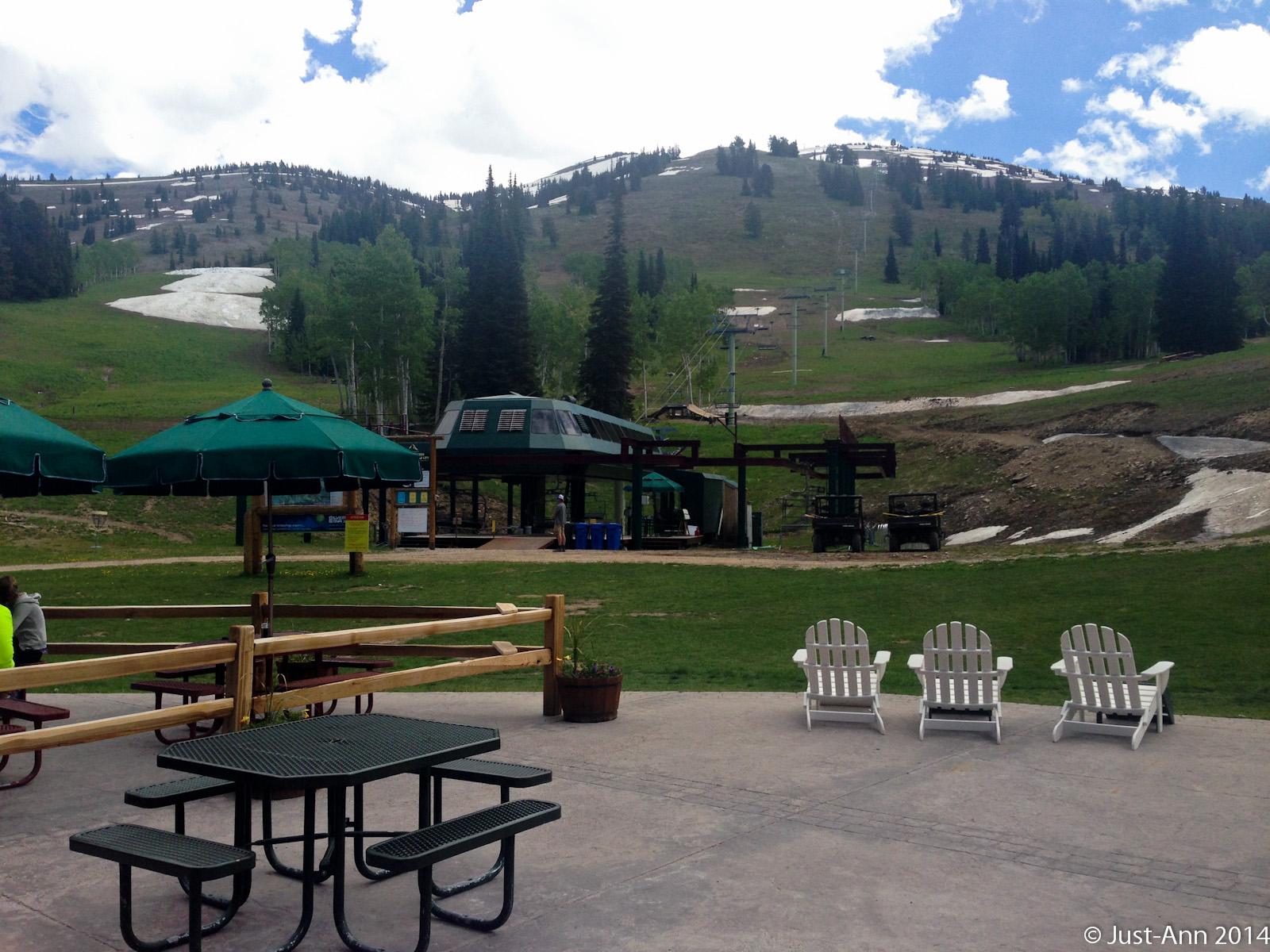 A scenic view of a mountain resort during springtime, featuring green grassy slopes, patches of snow, and a blue sky with scattered clouds. In the foreground, there are green picnic tables with umbrellas and white Adirondack chairs arranged in a row, alongside a ski lift station and grassy areas with trees in the background. Grand Targhee Bike Park mountain bike trail.