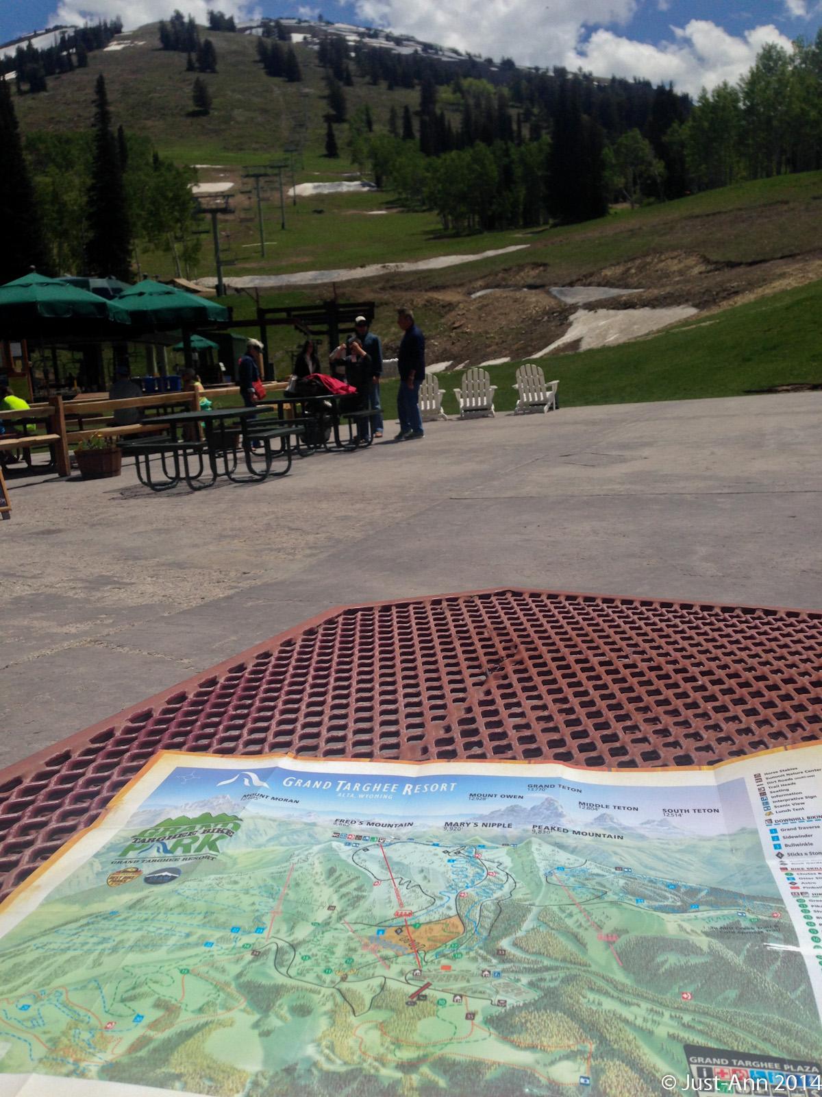 A map of Grand Targhee Resort is placed on a patterned picnic table, with a panoramic view of a ski hill in the background. The scene features green trees and a few patches of snow on the mountain, with people gathered in the foreground near outdoor seating areas. The sky is partly cloudy, indicating a pleasant day for outdoor activities. Grand Targhee Bike Park mountain bike trail.