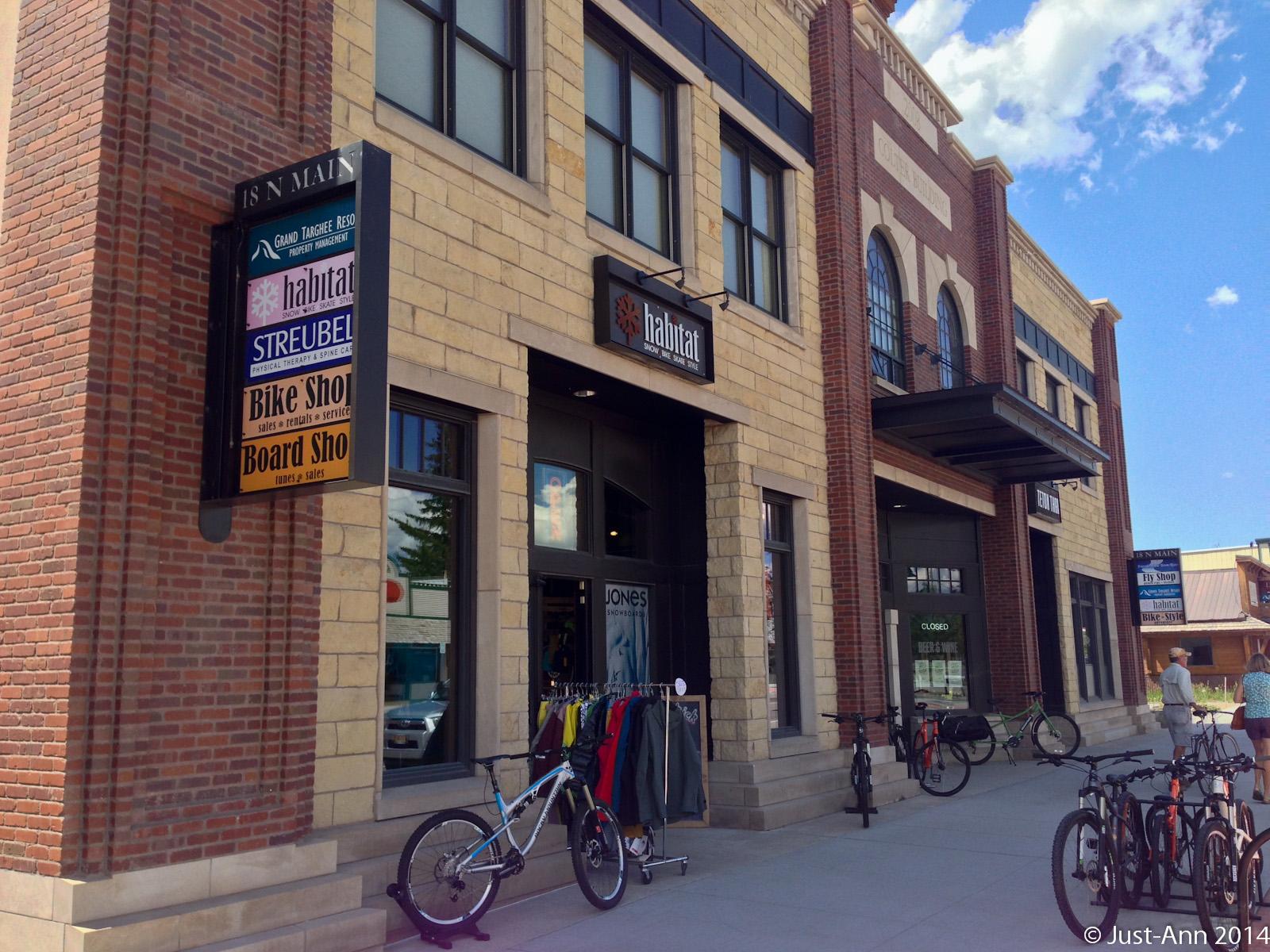 Exterior view of a brick building featuring multiple shops, including a bike shop and a snowboard shop. Bicycles are parked outside, and the sky is blue with scattered clouds. Signs for various businesses are visible, with a "CLOSED" sign on one of the doors.