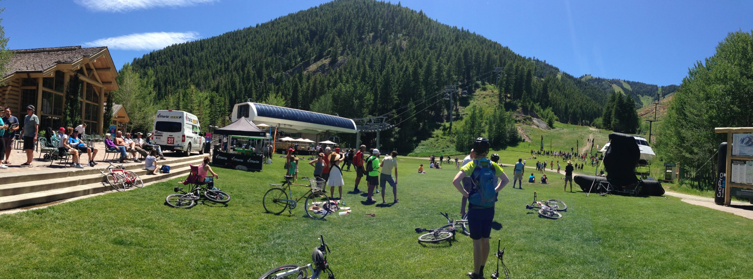 A panoramic view of a sunny day at a mountain biking event, featuring people mingling and relaxing on the grass. In the foreground, several cyclists stand near their bikes, while others are seated on the patio steps of a lodge. A vendor booth is set up nearby, and in the background, the green hills and trees are visible, along with a ski lift ascending the mountain. Bald Mountain Bike Park mountain bike trail.