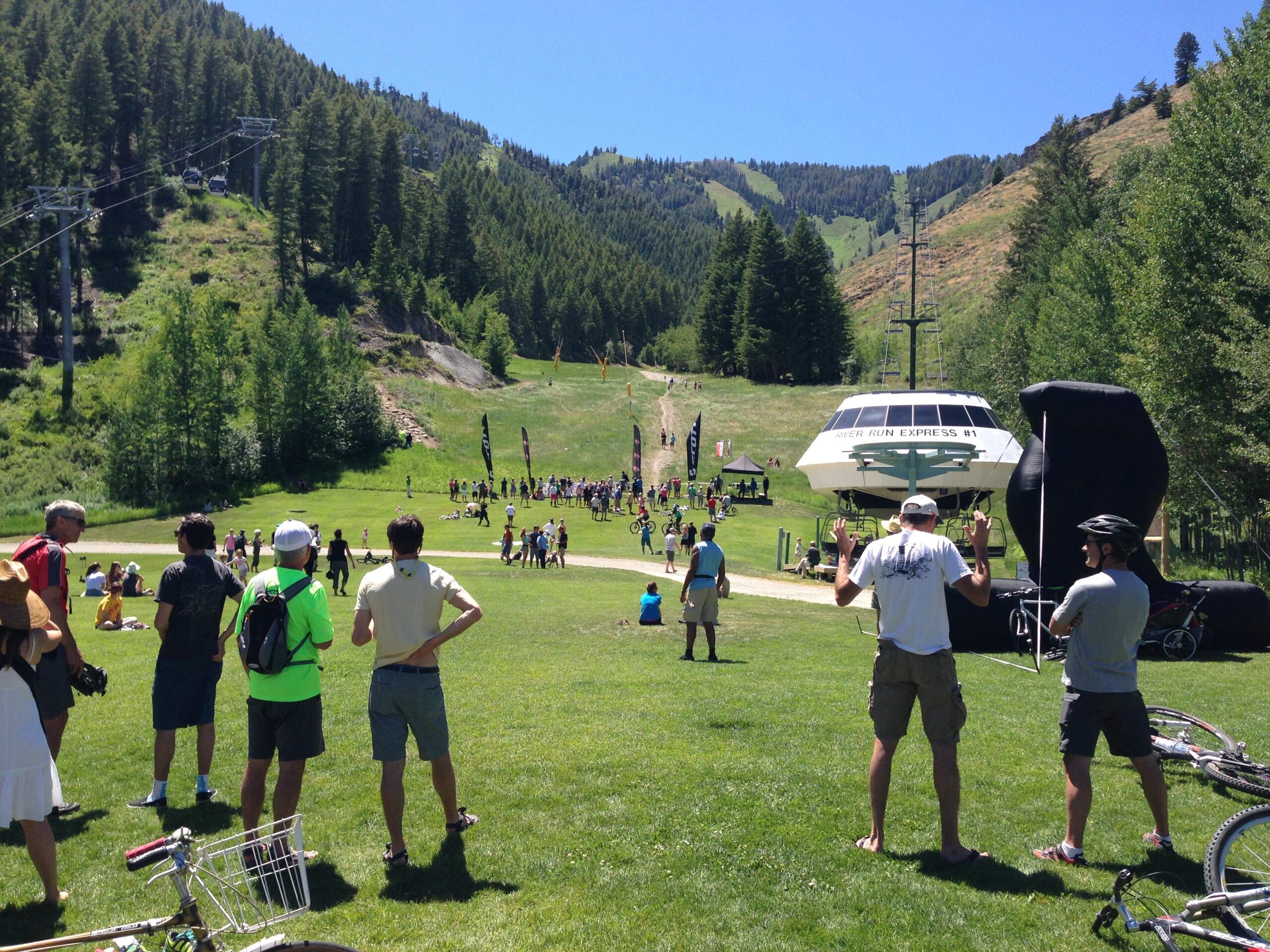 A scenic outdoor event at a mountain resort on a sunny day, featuring a crowd of people gathered on a green lawn. In the background, various participants are engaging in activities near a ski lift with a gondola visible. Some spectators are watching the event, while others are standing with bicycles nearby. Lush green trees and mountain slopes are visible in the backdrop. Bald Mountain Bike Park mountain bike trail.