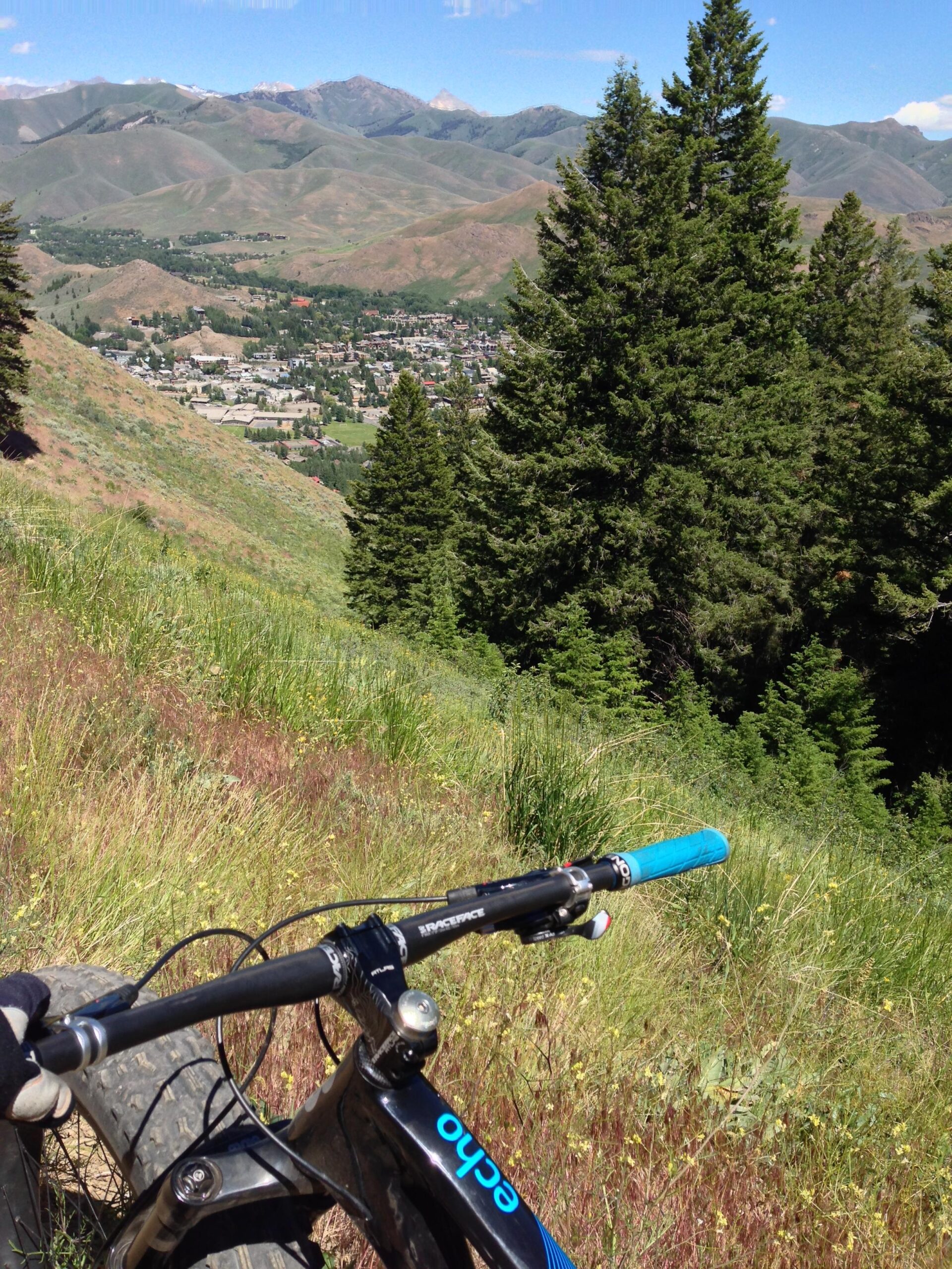 Mountain bike handlebars in the foreground with a scenic view of a valley and town below, surrounded by rolling hills and mountains under a clear blue sky. Dense green trees are visible on the hillside. Bald Mountain Trail mountain bike trail.