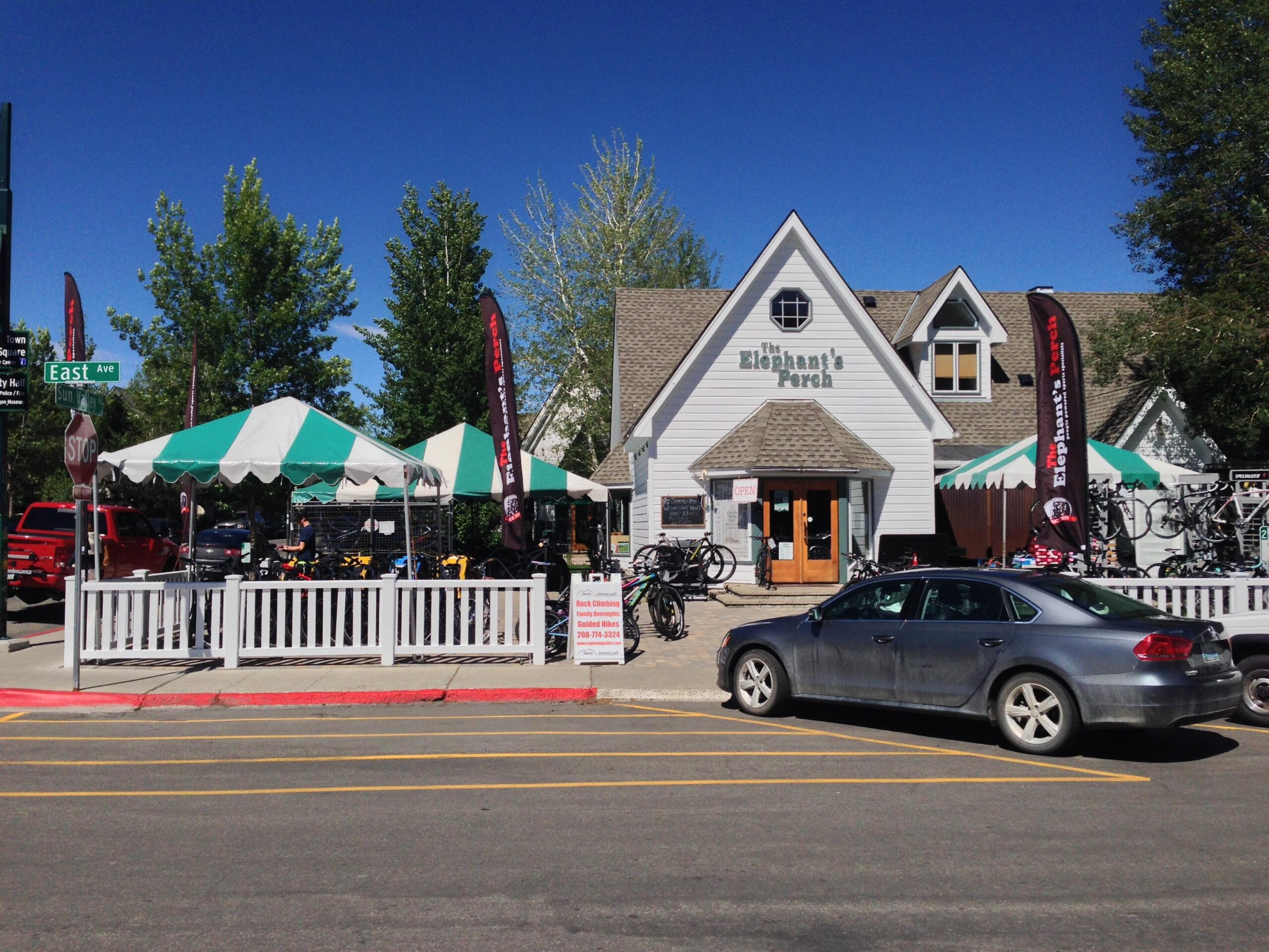 Exterior view of "The Elephant's Porch," a bike shop with a white and gray building and a pointed roof. The shop features green and white striped tents outside, displaying various bicycles for sale. A few trees provide greenery in the background, and a parked car is in the foreground, with a street sign indicating "East Ave" nearby. The sky is clear and blue, suggesting a sunny day.