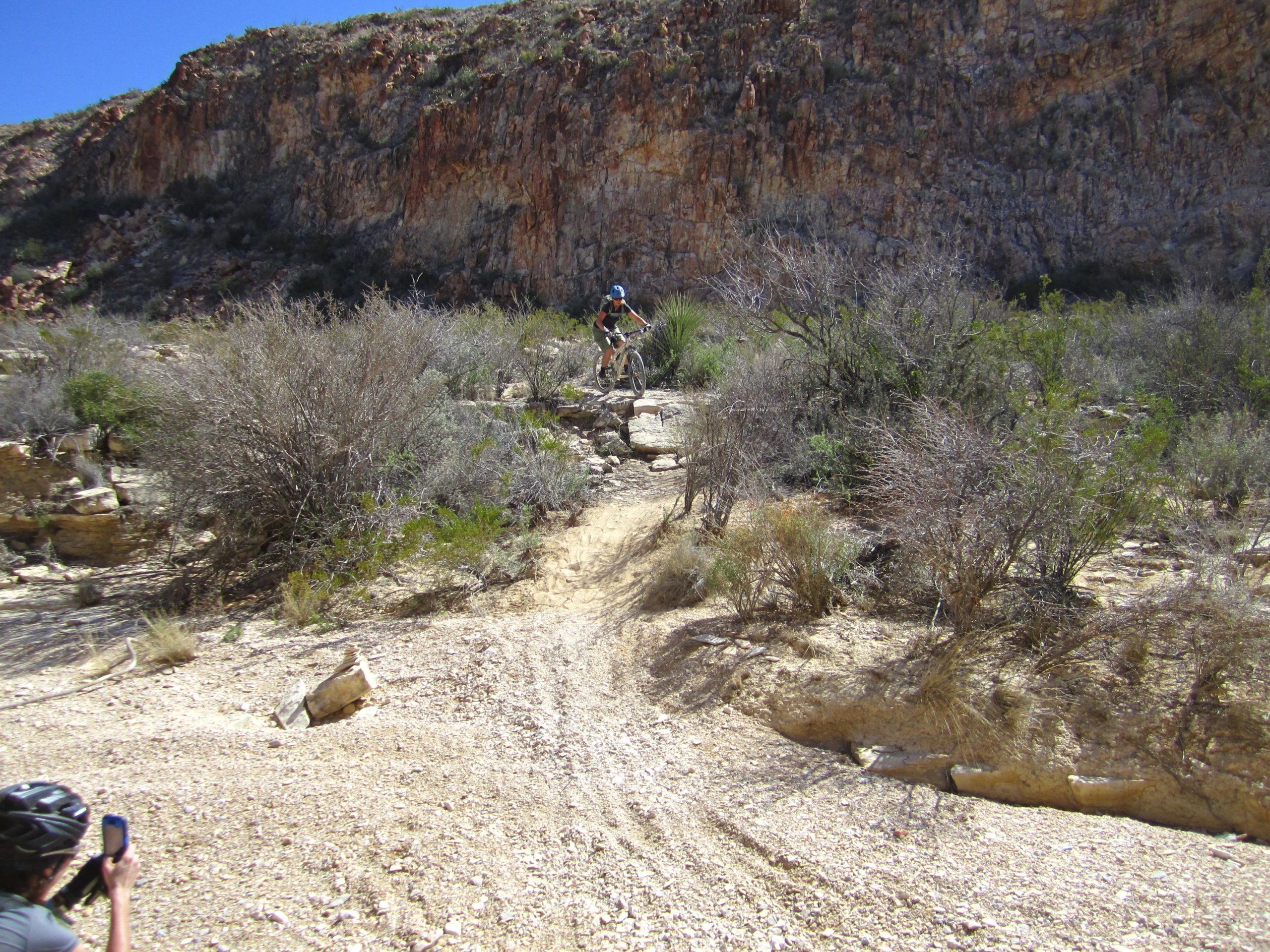A mountain biker navigating a rocky trail in a desert landscape, with steep cliffs in the background and sparse vegetation surrounding the path. Another person in the foreground is taking a photo of the biker. Big Bend Ranch State Park mountain bike trail.