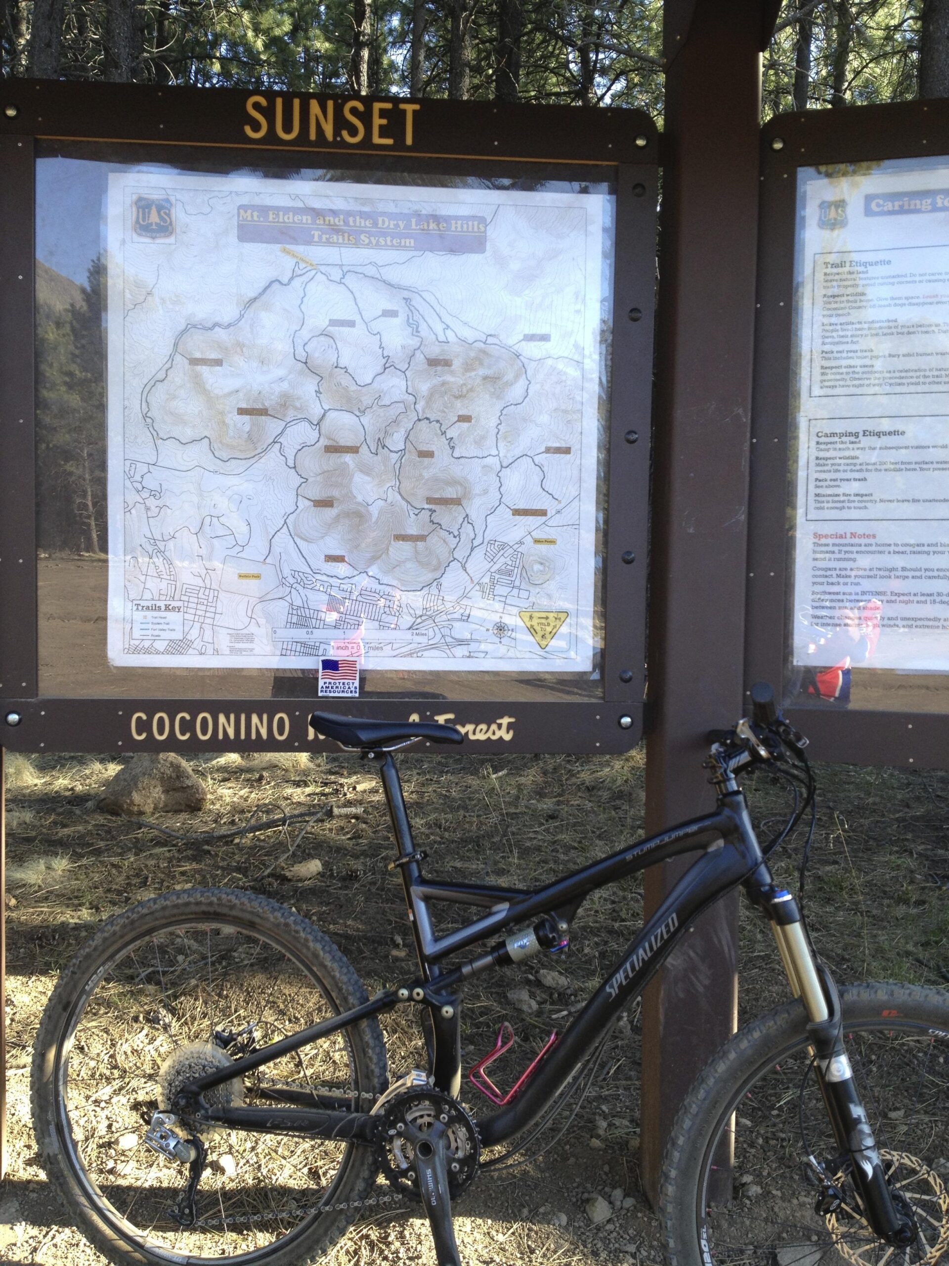Specialized Stumpjumper FSR: A mountain bike leaning against a trail map sign for the Mt. Elden and the Dry Lake Hills Trails System, surrounded by trees in a forested area. The sign includes trail information and guidelines, with "SUNSET" prominently displayed at the top.
