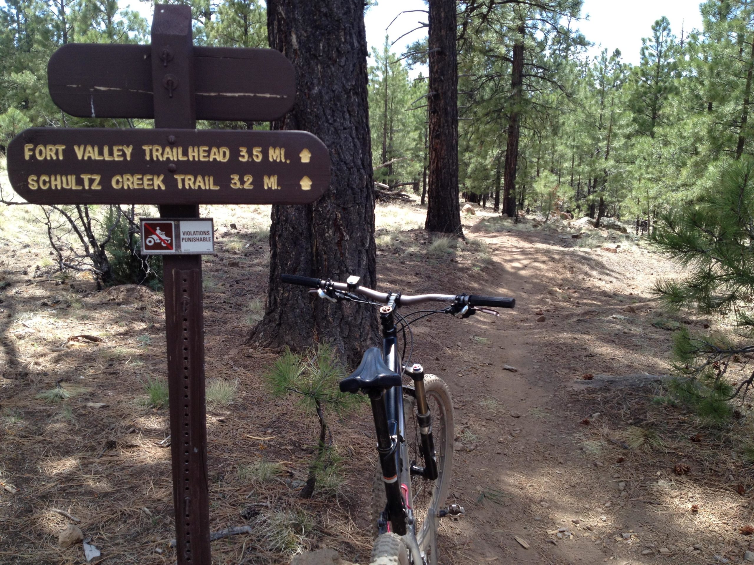 A mountain bike rests beside a wooden trail sign indicating directions and distances to Fort Valley Trailhead (3.5 miles) and Schultz Creek Trail (3.2 miles) in a wooded area with pine trees and a dirt path. Fort Valley Trail System mountain bike trail.