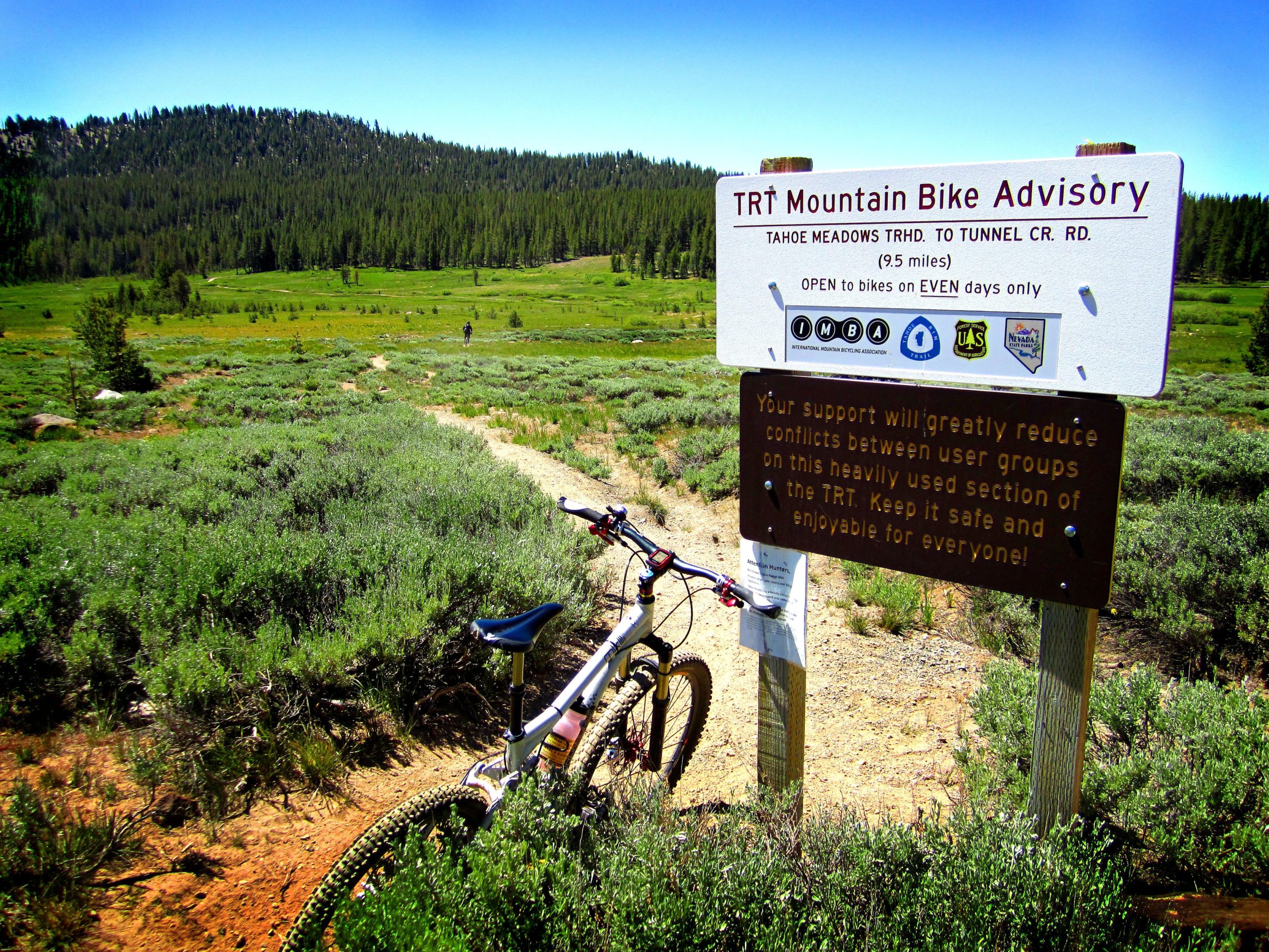 A mountain bike rests on a dirt path surrounded by green vegetation. In the background, a sign reads "TRT Mountain Bike Advisory," providing information about the Tahoe Meadows to Tunnel Creek Road trail. A hiker is visible in the distance, and the scene is set against a backdrop of evergreen trees under a clear blue sky. Tahoe Rim Trail: Tahoe Meadows to Tunnel Creek Road / Flume Trail mountain bike trail.
