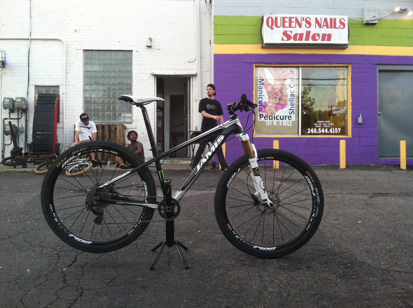 Jamis Nemesis: A black and white mountain bike stands on a stand in front of a colorful salon with a purple and green exterior. In the background, two people are seated and another is standing, while a small delivery cart is visible next to the building. The scene is set on a pavement with some worn areas.