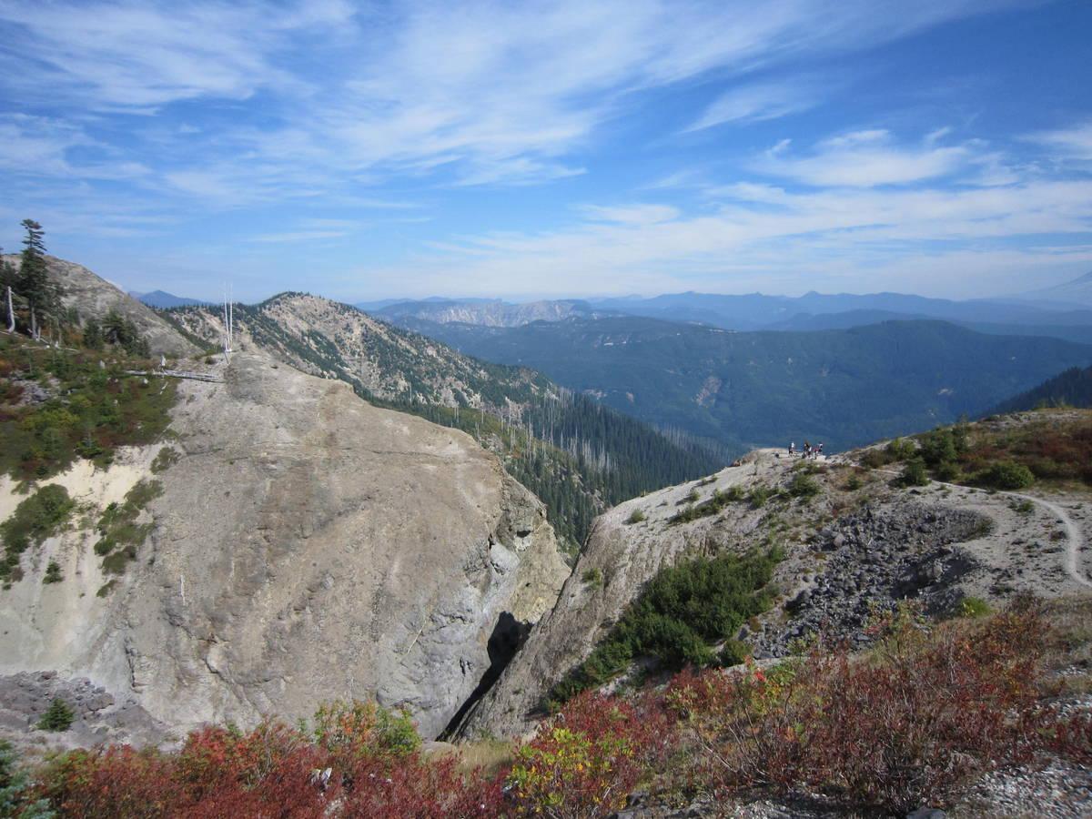 A panoramic view of a mountainous landscape, featuring rugged cliffs and green forested slopes under a blue sky with wispy clouds. In the distance, layers of mountains fade into the horizon. A small group of hikers can be seen on a rocky outcrop, surrounded by vibrant autumn foliage in the foreground. Ape Canyon#234, Abraham#216d, Smith Creek#225 Trails mountain bike trail.