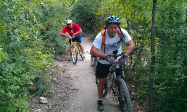 Two cyclists on a mountain biking trail surrounded by greenery. One cyclist, wearing a white shirt and black shorts, pushes his bike up the path, while another cyclist in a red shirt and yellow bike rides ahead. The trail is narrow and winding, with trees and bushes lining the route. Sansom Park mountain bike trail.