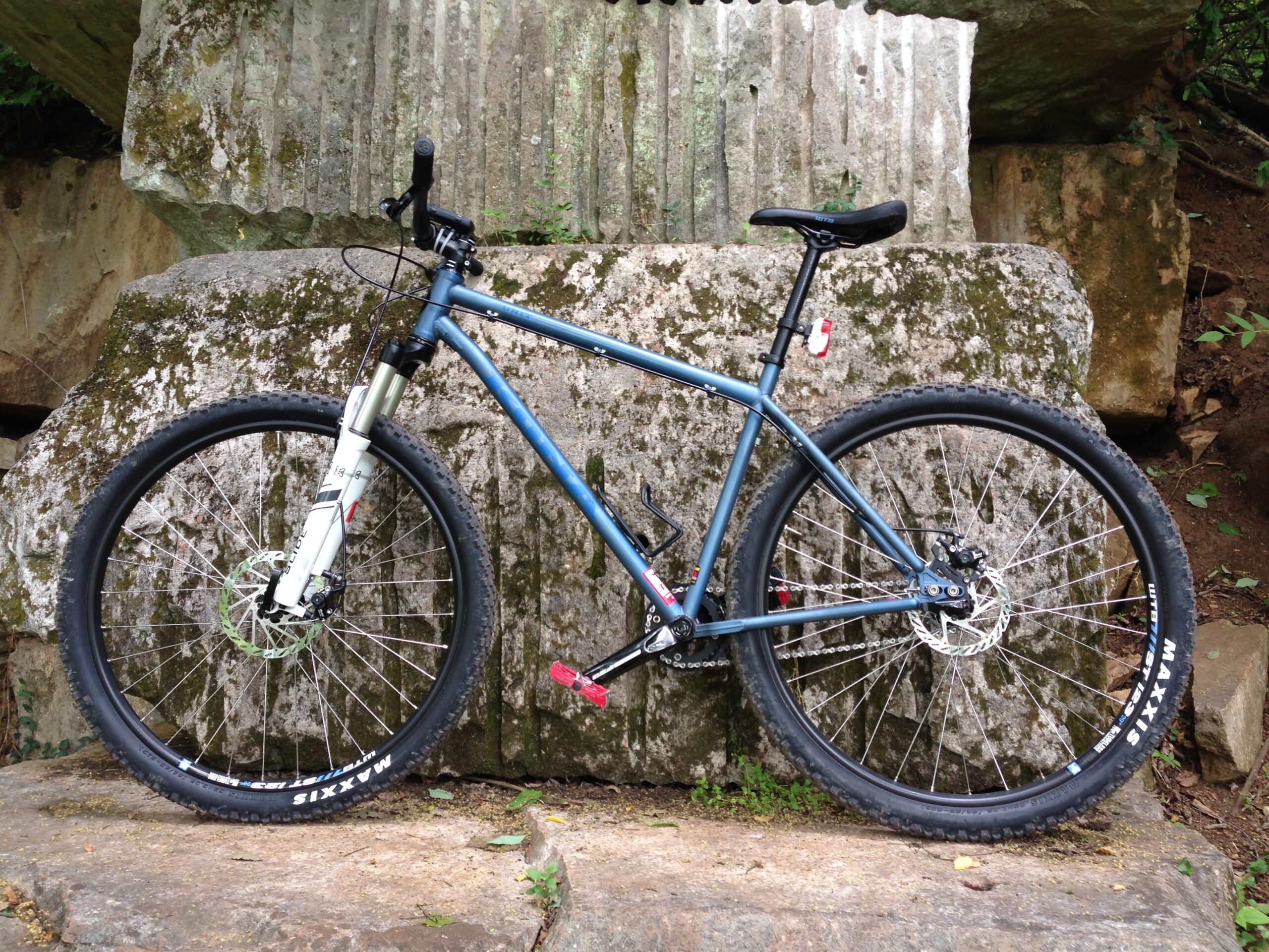 Kona Unit: A mountain bike leaning against a large rock with a textured surface, surrounded by greenery. The bike features a blue frame, thick tires with Maxxis branding, and a white front suspension fork.