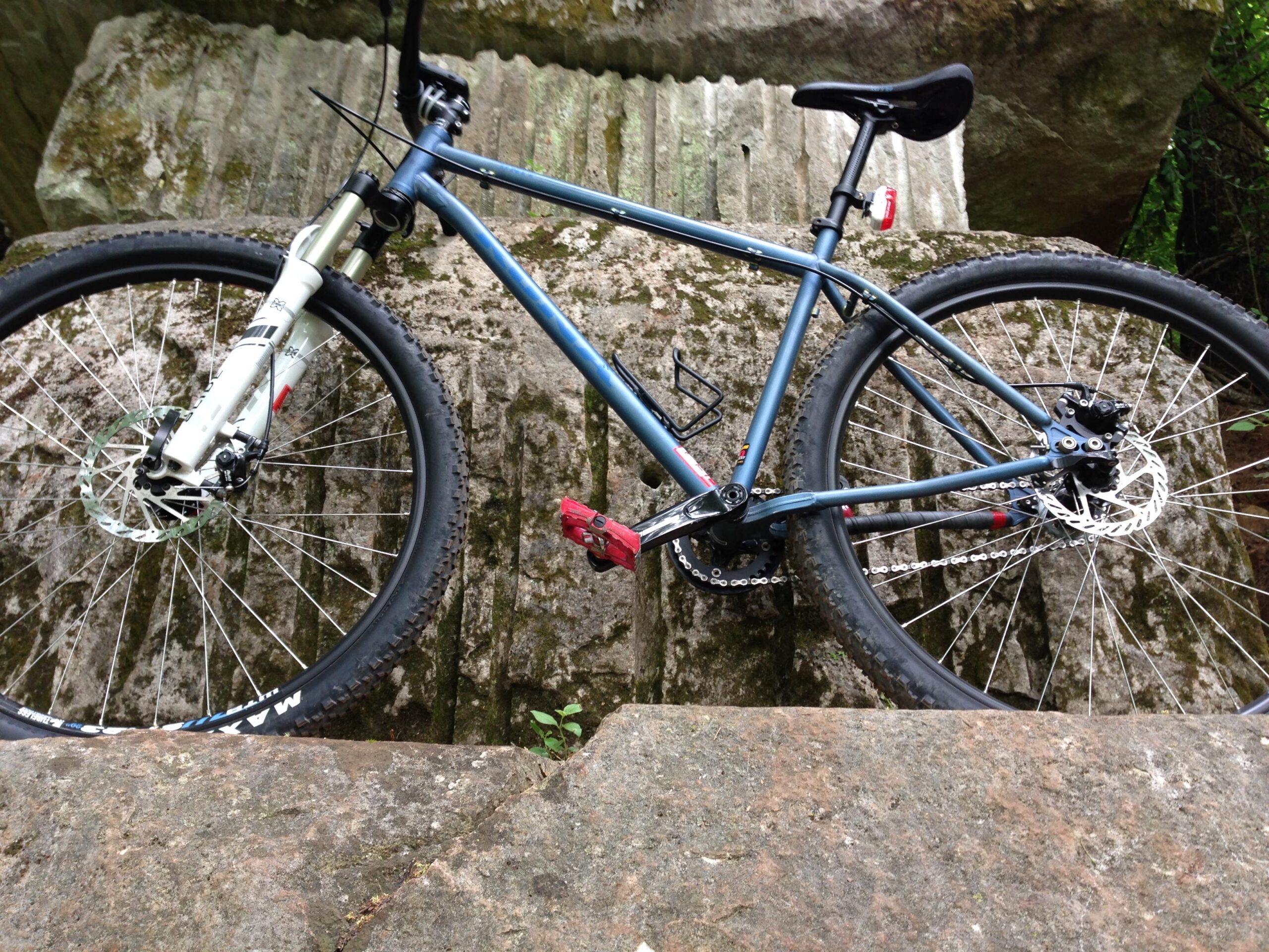 Kona Unit: A blue mountain bike positioned on a rocky surface, featuring thick tires, disc brakes, and a suspension fork, with greenery in the background.