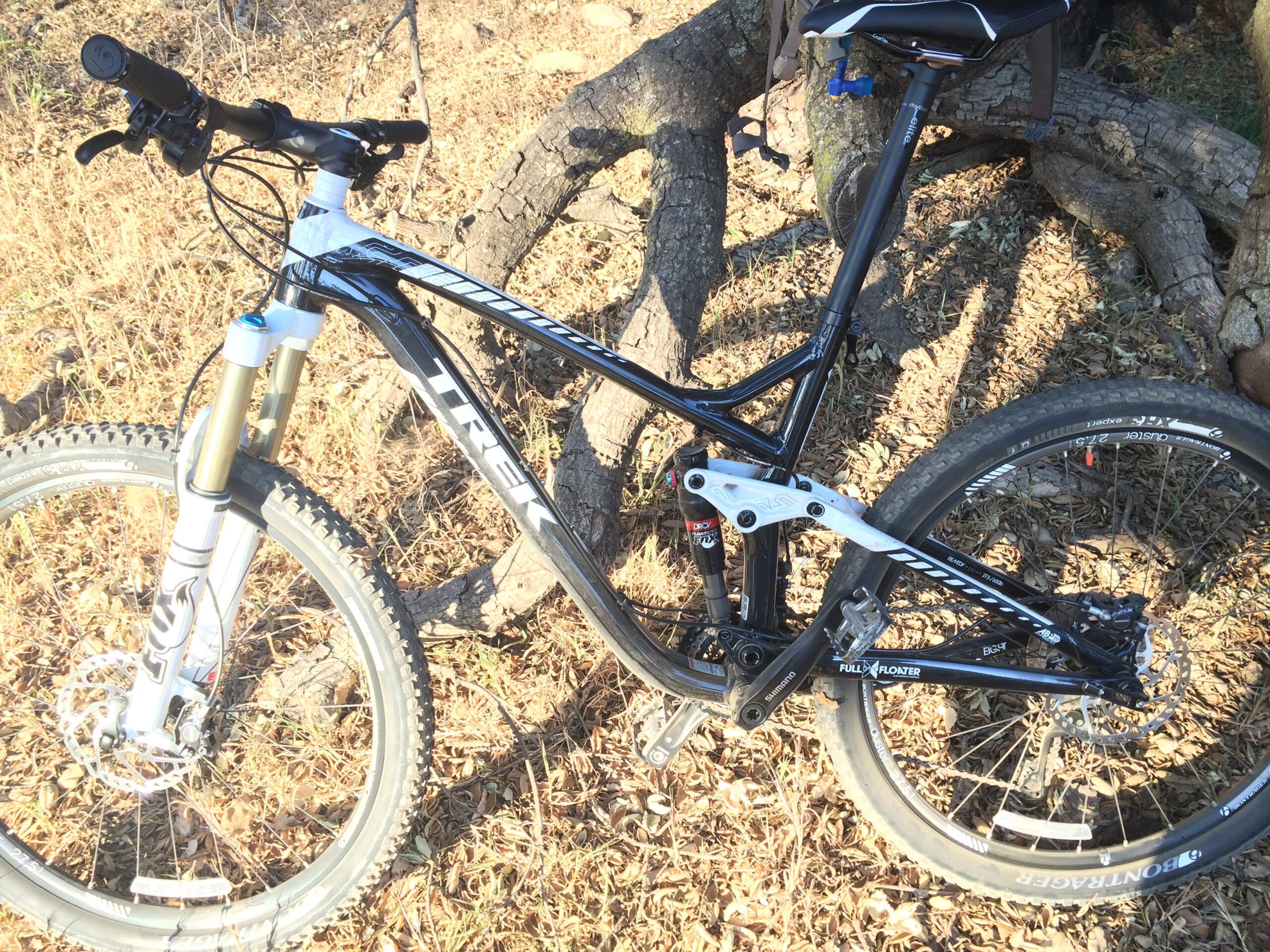 Trek Remedy 8: A black Trek mountain bike leaning against a tree, featuring a front fork with suspension, knobby tires, and a minimalist design. The bike is positioned on a bed of dry leaves, with sunlight casting shadows on the ground.
