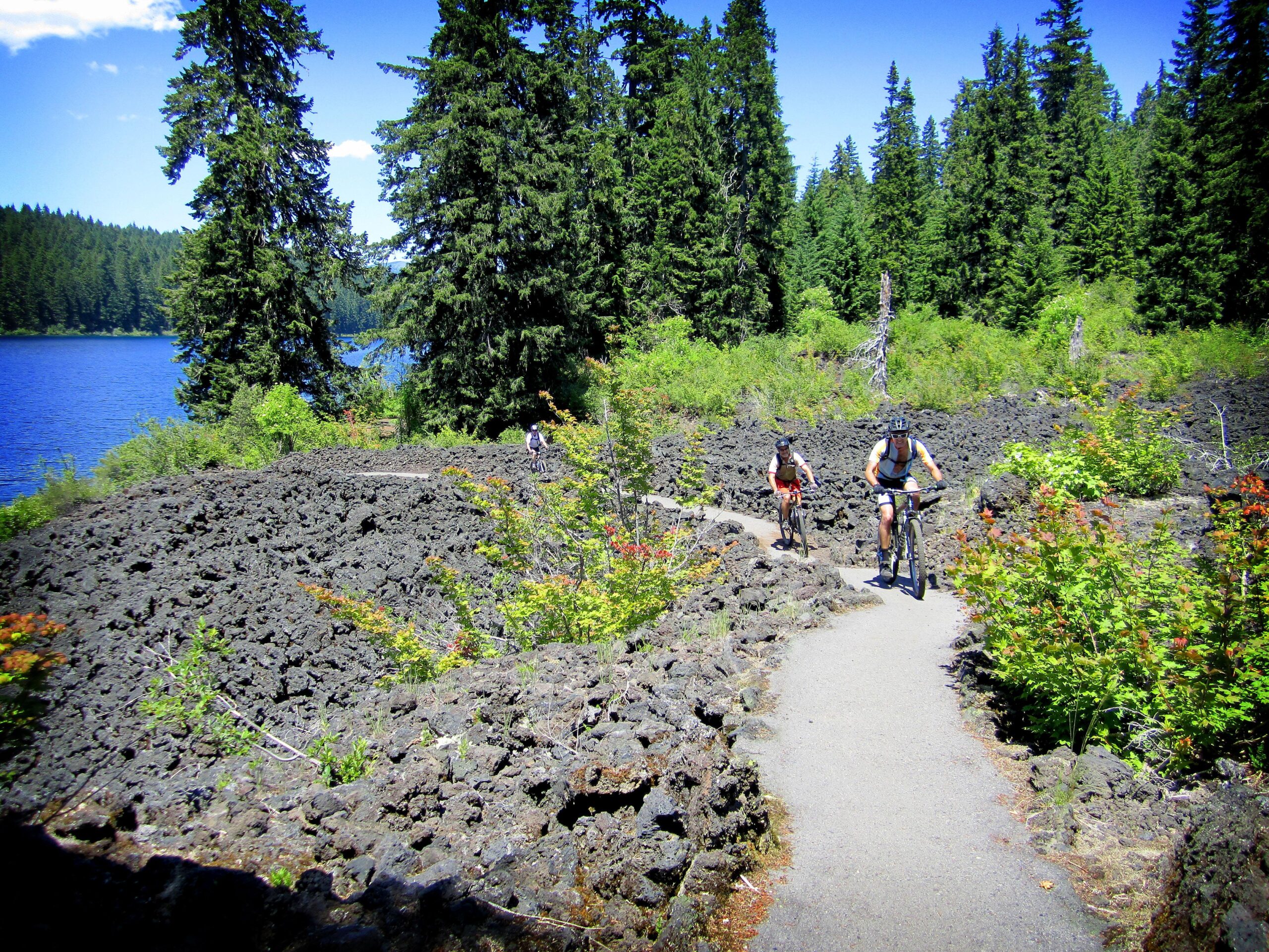 A scenic outdoor landscape featuring two mountain bikers riding along a winding path through a rocky, volcanic terrain. In the background, a blue lake is visible, surrounded by lush green trees and dense vegetation under a clear blue sky. The setting is vibrant and inviting for outdoor activities. Mckenzie River Trail mountain bike trail.