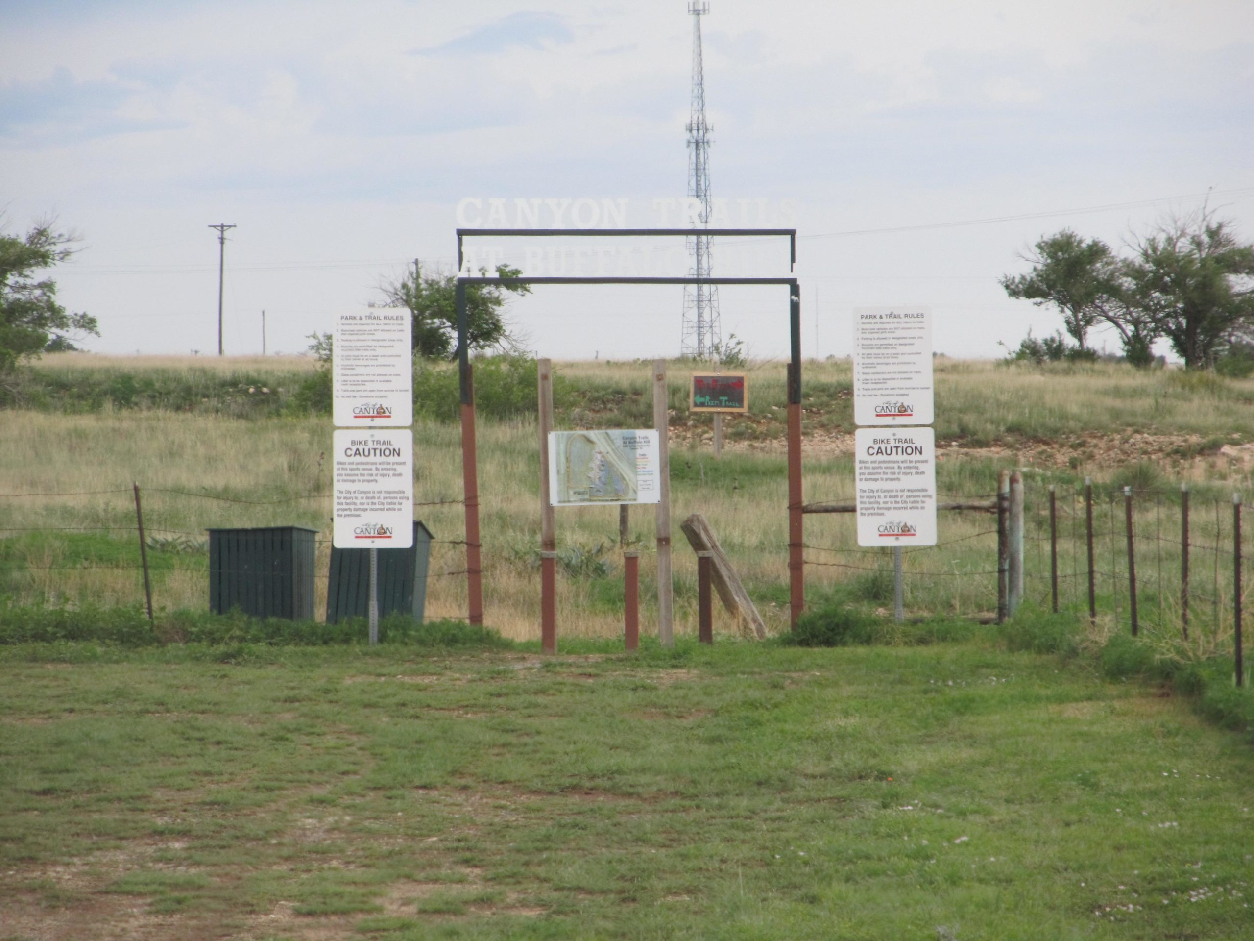 A wooden entrance sign for Canyon Trails at Buffalo with multiple informational signs about park and trail rules, including a caution notice for bike trails. The background features grassy fields and a distant cell tower. Buffalo Trails mountain bike trail.