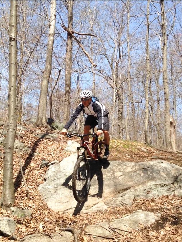 A mountain biker navigating over a large rock on a trail in a forest. Surrounding trees are bare, indicating early spring, with scattered leaves on the ground. The cyclist is wearing a helmet and a jacket, focused on balancing and maneuvering the bike. Ramapo Mountain State Forest mountain bike trail.