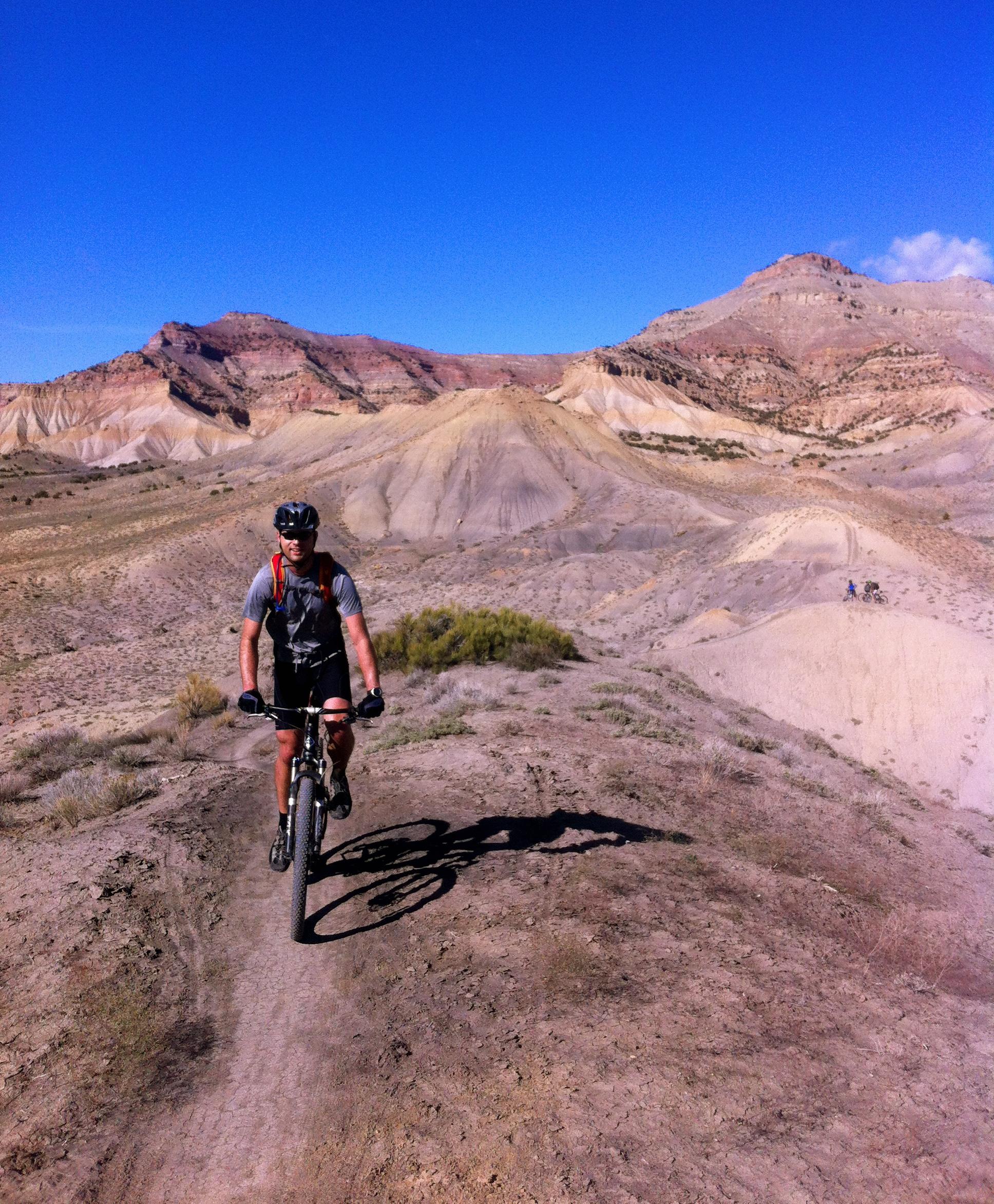 A mountain biker riding along a dirt trail in a rugged, desert landscape with colorful hills and a clear blue sky in the background. 18 Road Trails / North Fruita Desert mountain bike trail.