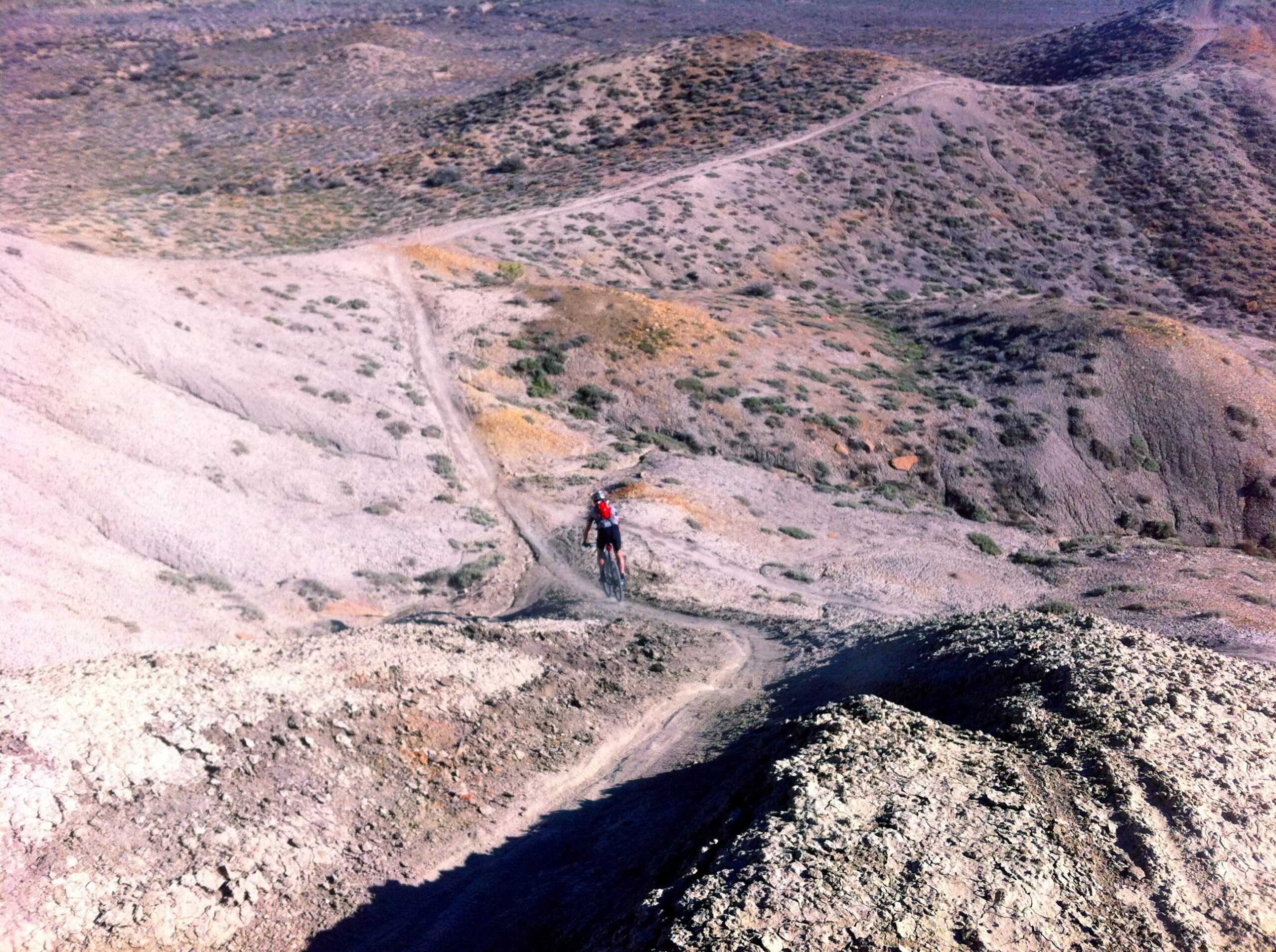 A cyclist rides along a winding dirt path on a hilly terrain, surrounded by sparse vegetation and rocky landscapes under a clear sky. The view showcases the expansive, rugged landscape with rolling hills and a prominent path leading into the distance. 18 Road Trails / North Fruita Desert mountain bike trail.