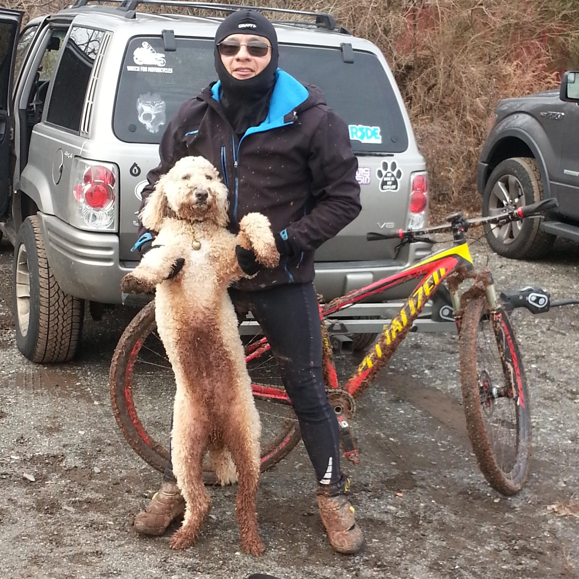 A person wearing cycling gear and sunglasses is holding a muddy dog in a playful pose. The background features a parked car and a mountain bike leaning against it, both covered in mud. The scene captures a fun outdoor moment after a ride. Six Mile Run mountain bike trail.