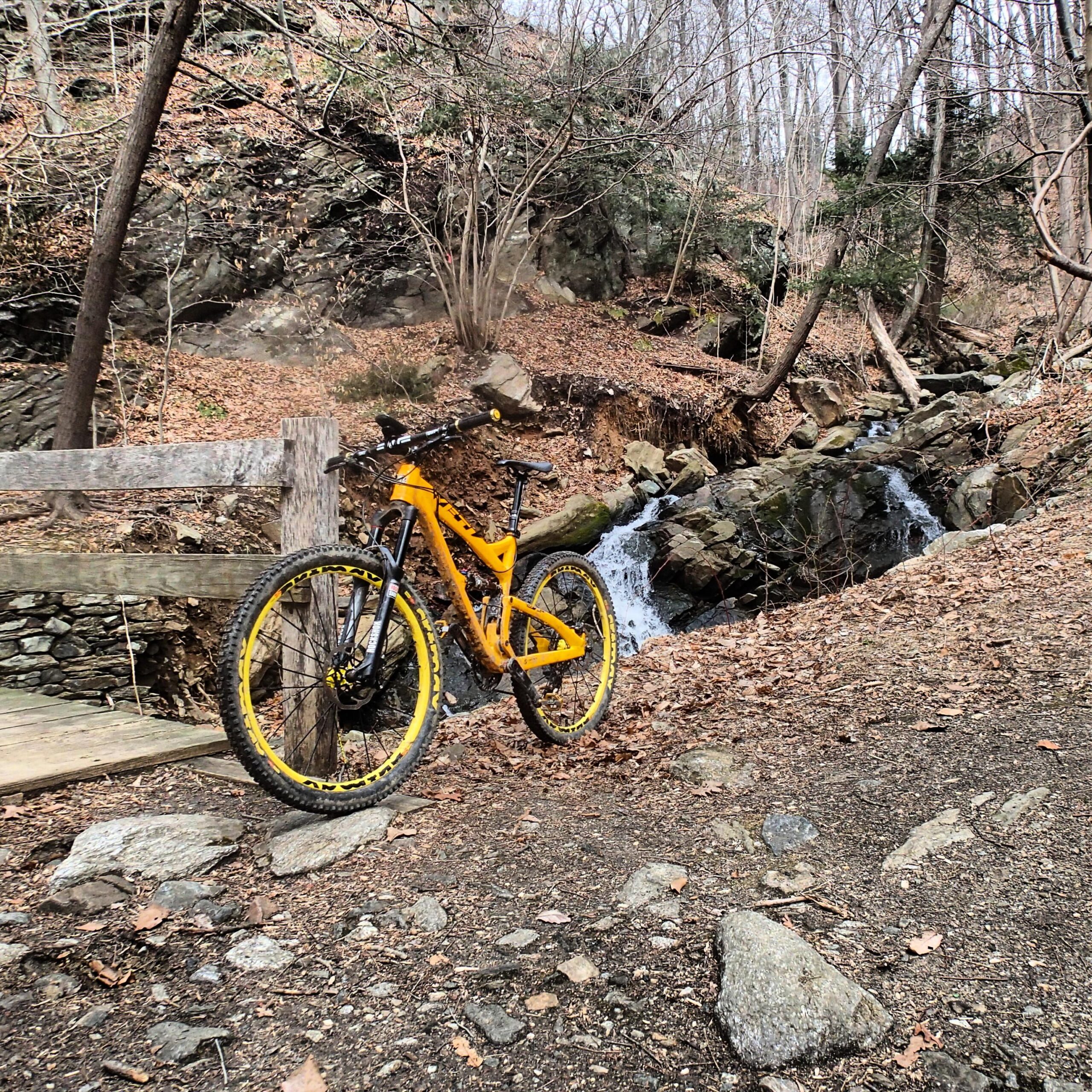 A yellow mountain bike rests near a small waterfall, surrounded by rocky terrain and bare trees. Fallen leaves cover the ground, and a wooden fence and footbridge are visible in the background. Wissahickon Valley Park mountain bike trail.