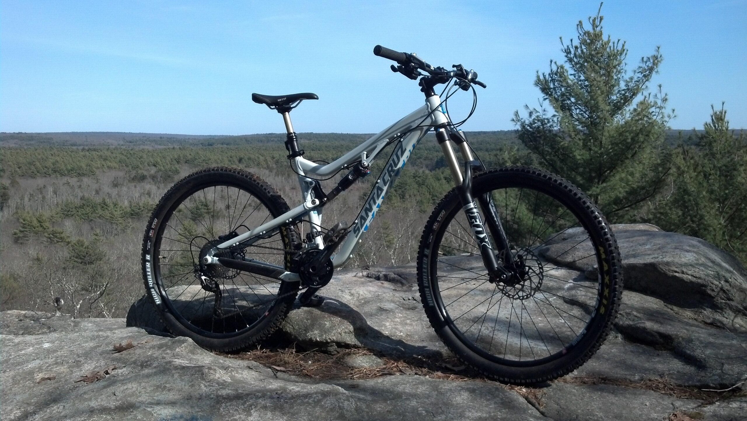 Santa Cruz Bronson: A mountain bike resting on a rocky outcrop, with a scenic view of a forested landscape in the background. The sky is clear and blue, showcasing a perfect day for outdoor activities.