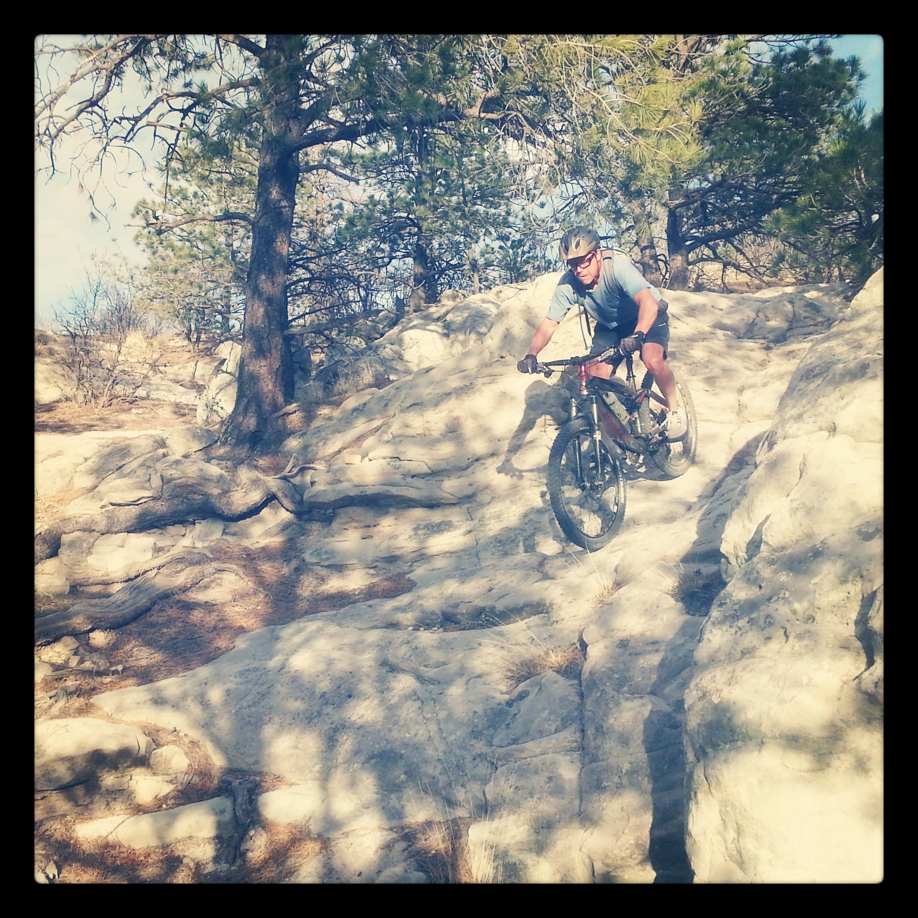 A mountain biker navigating a rocky trail surrounded by trees and blue sky, showing concentration and skill as he rides over rugged terrain. Ute Valley Park mountain bike trail.