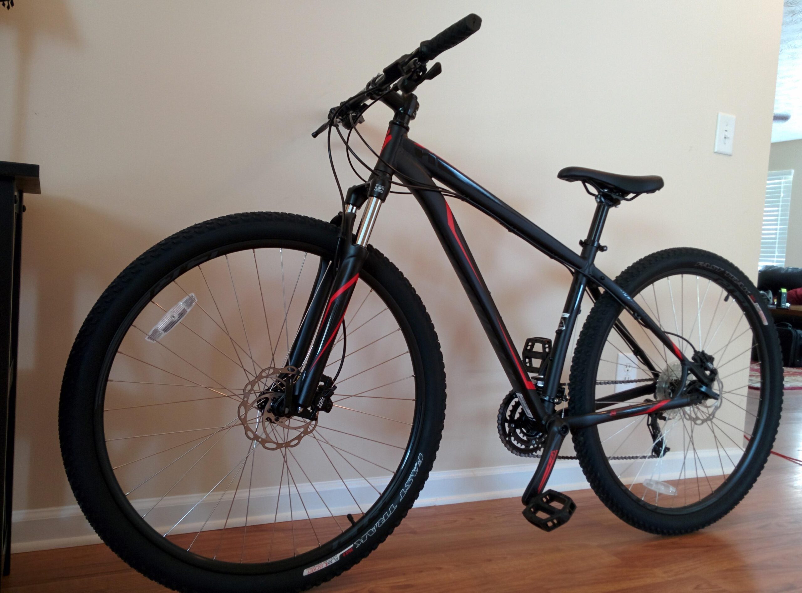 Specialized Hardrock Sport Disc 29er: A black and red mountain bike with thick tires, disc brakes, and a sleek frame, positioned against a light-colored wall. The bike features a comfortable seat and flat pedals, showcasing its design for off-road cycling.