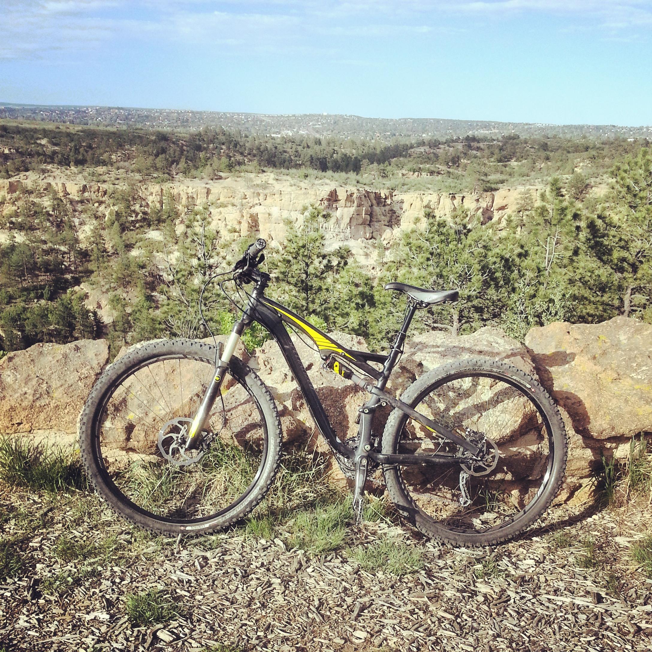 A mountain bike resting on a rocky surface with a scenic backdrop of trees and hills under a blue sky. Palmer Park mountain bike trail.