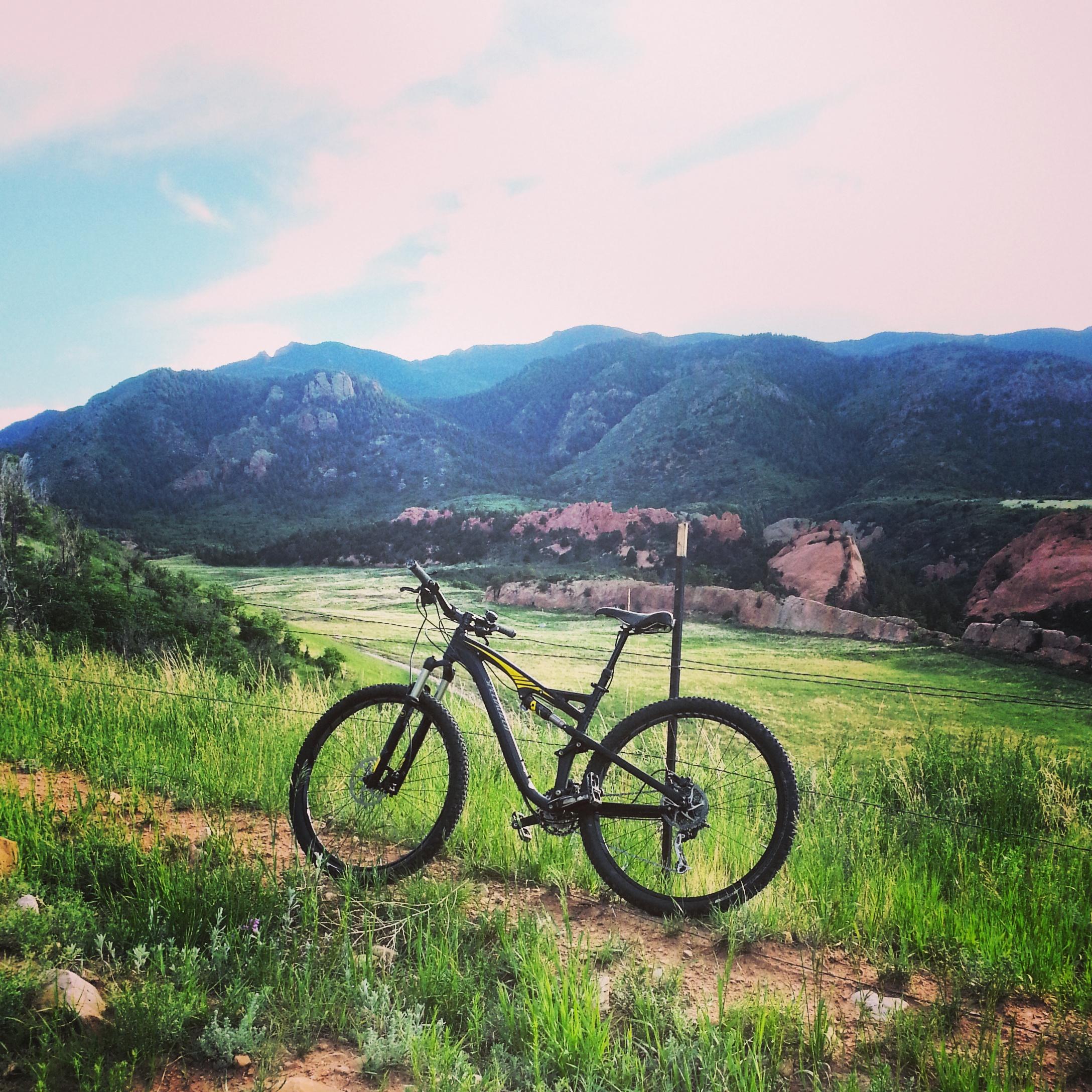 A mountain bike parked on a dirt path with a scenic view of green hills and rocky mountains in the background under a clear blue sky. Red Rock Canyon mountain bike trail.