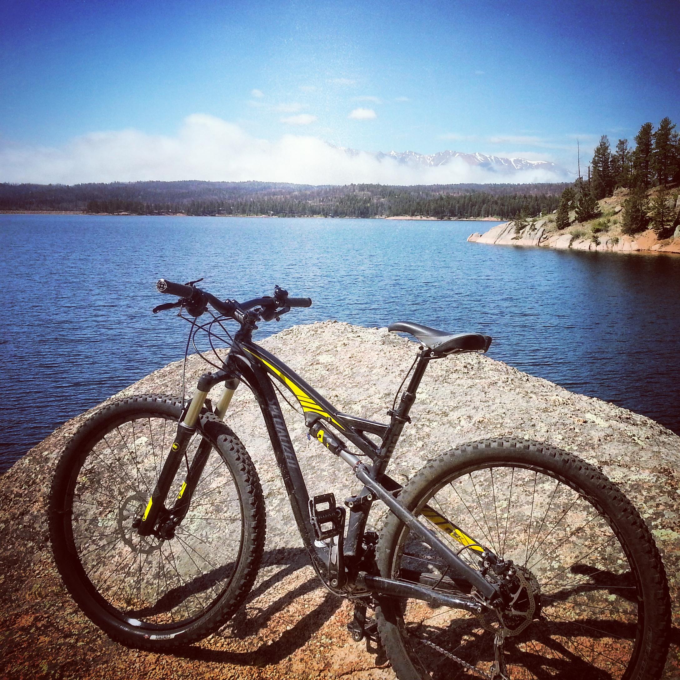 A mountain bike rests on a rocky ledge overlooking a tranquil lake, with a backdrop of trees and distant mountains under a clear blue sky. Rampart Reservoir mountain bike trail.