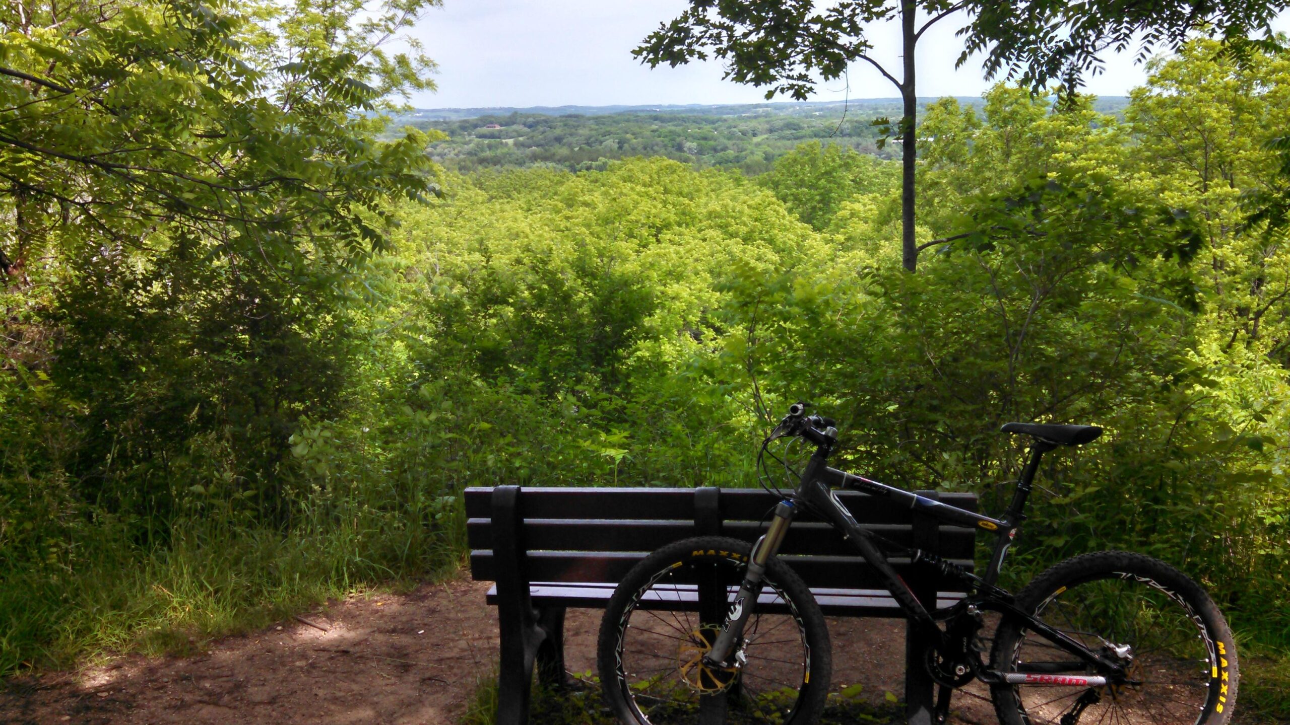 A black mountain bike is parked next to a wooden bench overlooking a lush green landscape, with trees and hills in the distance under a partly cloudy sky. Emma Carlin mountain bike trail.