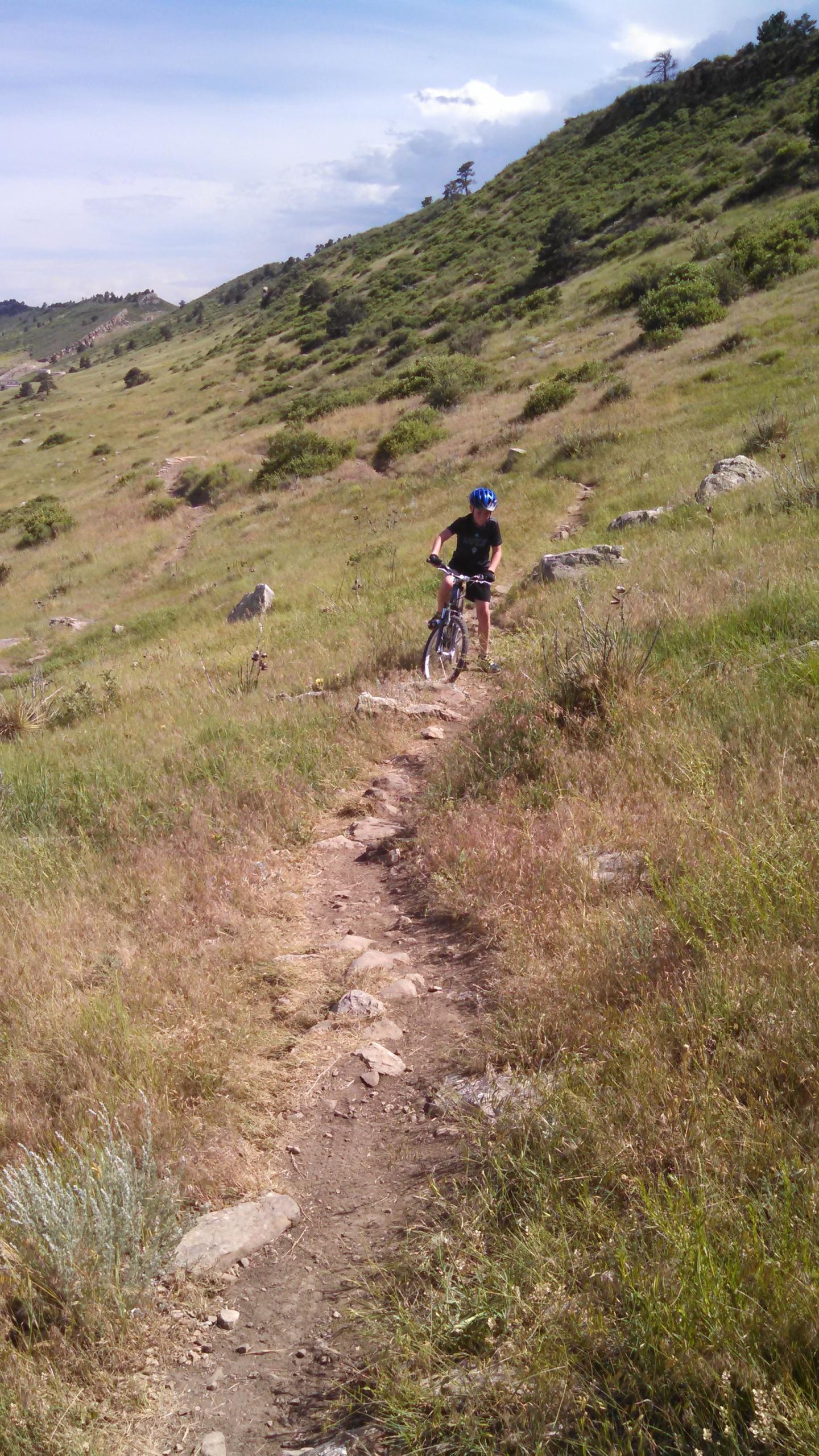 A young person in a blue helmet stands next to a mountain bike on a rocky path, surrounded by tall grass and shrubs on a hillside. The background features rolling hills and a partly cloudy sky. Reservoir Ridge mountain bike trail.