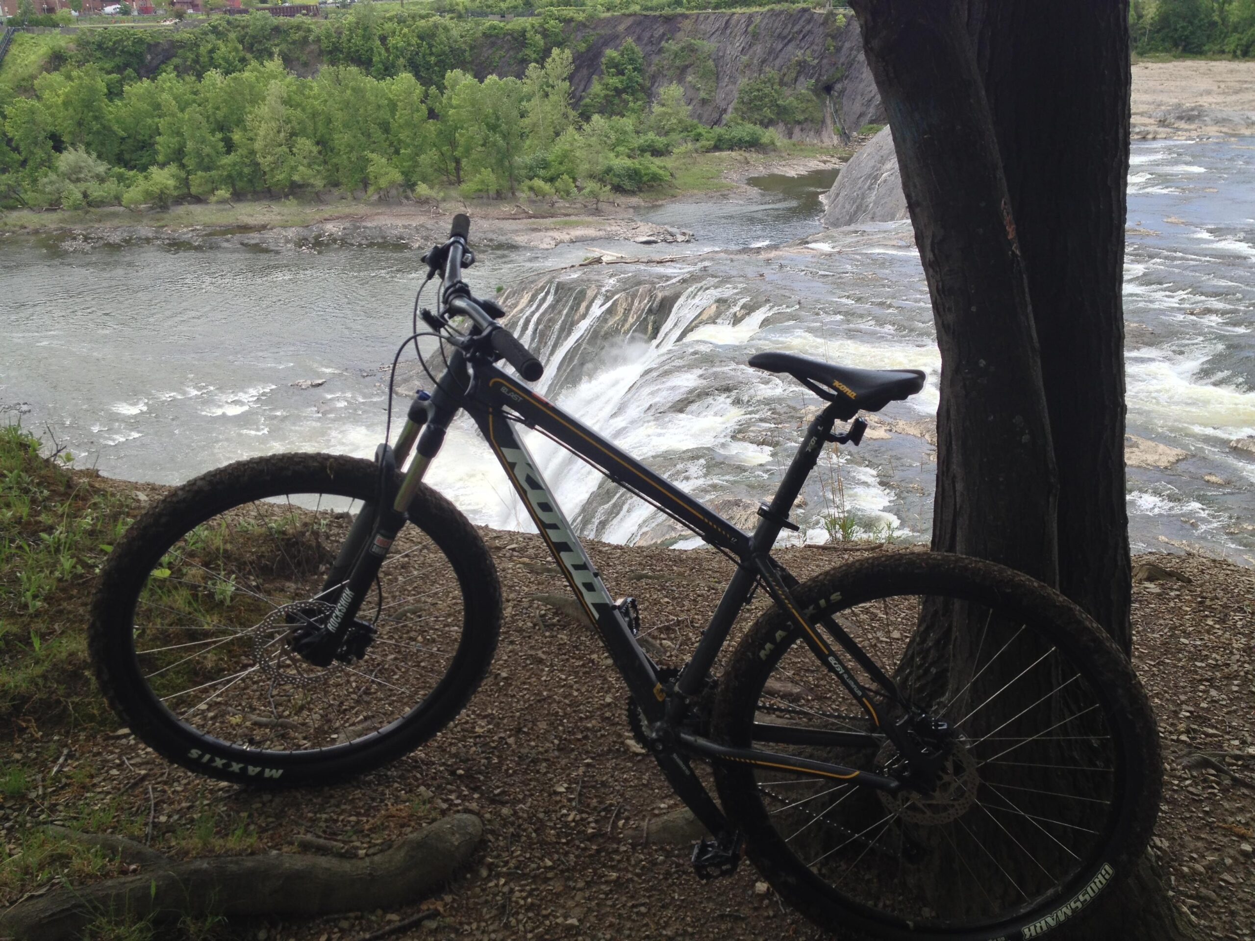 Kona Blast: A mountain bike resting against a tree, overlooking a river with a waterfall casc situate in a lush green landscape. The scene captures the contrast between the bike and the natural surroundings, depicting a tranquil outdoor adventure setting.