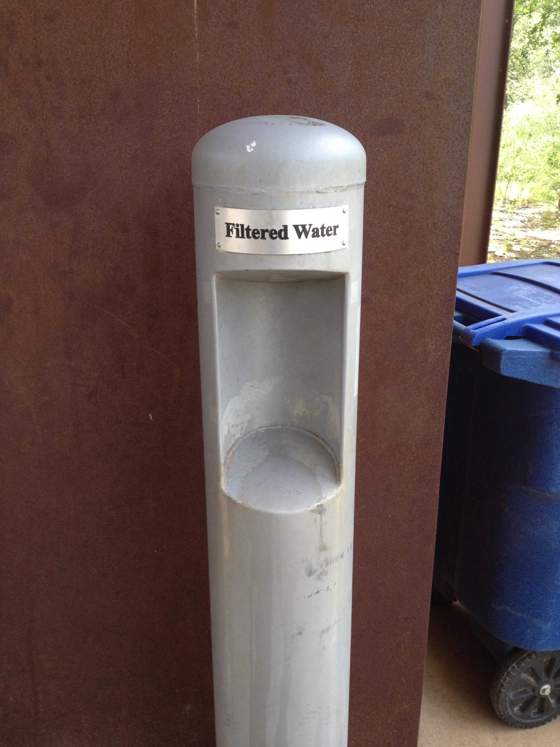 A vertical drinking fountain labeled "Filtered Water," made of metal with a rounded top, positioned next to a blue trash bin. The background features a textured brown surface. Salado Creek mountain bike trail.