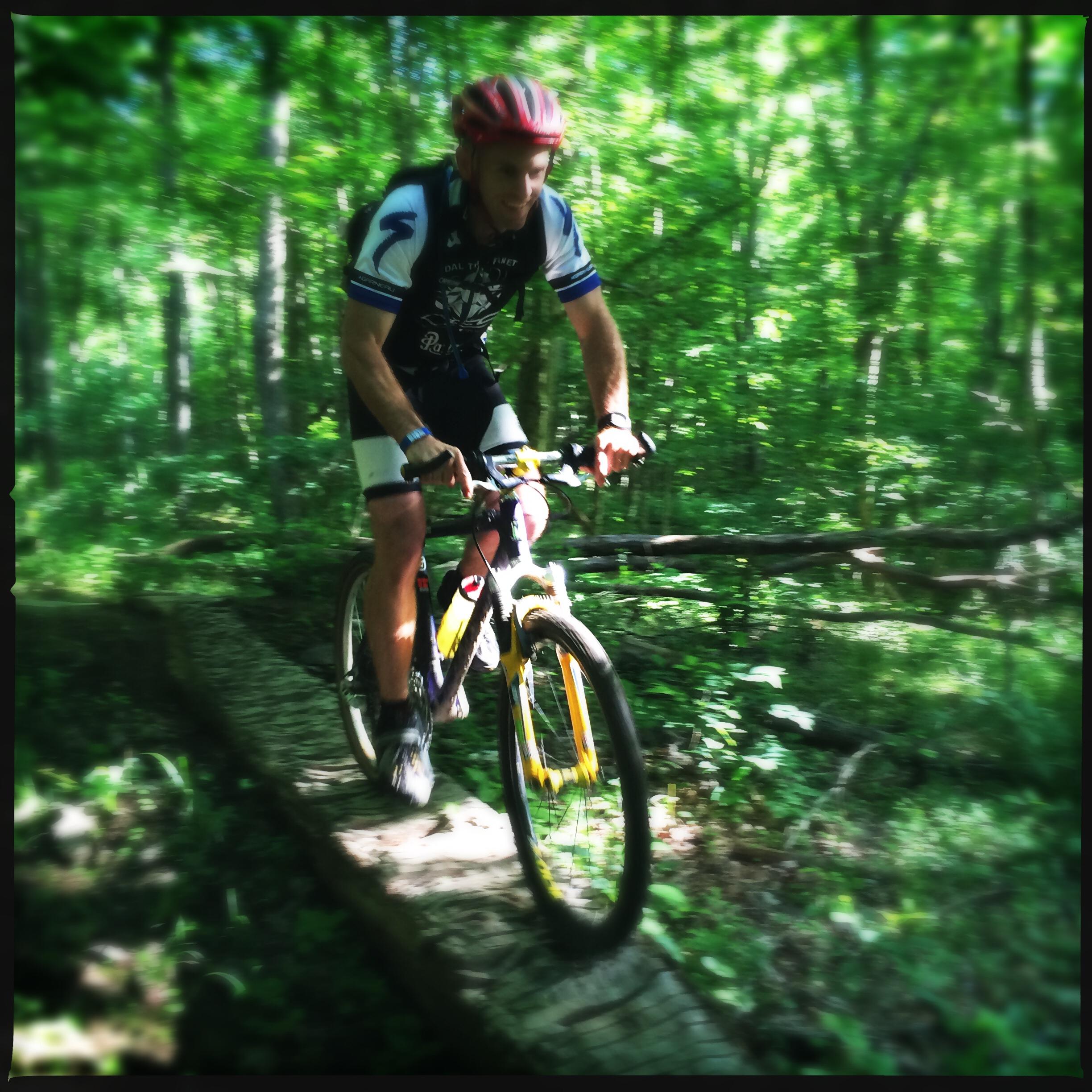 A mountain biker in a helmet rides a yellow bike across a narrow wooden bridge in a lush, green forest. The scene captures the motion and focus of the cyclist as sunlight filters through the trees, creating a vibrant and dynamic atmosphere. Skullbuster mountain bike trail.